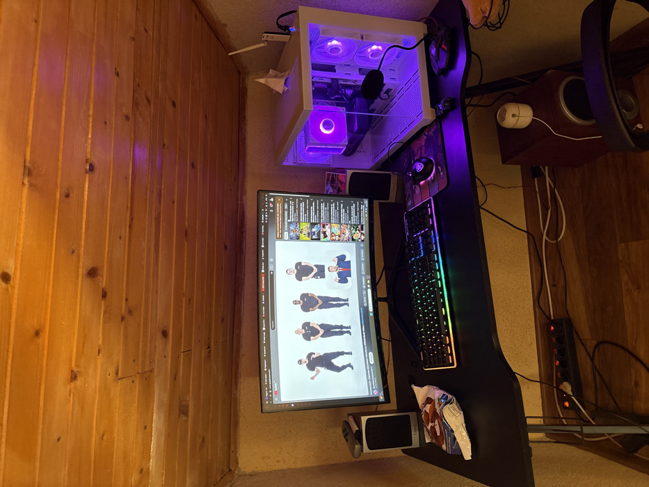 The image depicts a gaming setup within an indoor room located in the city of Stefan Karadja, Bulgaria. The room features a low, sloped ceiling constructed from horizontal wooden planks and textured walls painted a light, warm hue. The floor is covered with a wood-patterned laminate.
A black L-shaped gaming desk occupies the foreground, supporting a large, curved monitor that displays a YouTube video. The video shows five male subjects: four in black t-shirts and black pants standing in a line, and a fifth subject, resembling Donald Trump, in a suit and red tie, giving a thumbs-up gesture below them. To the right of the monitor, a white computer tower with a transparent side panel is internally illuminated by purple LED lights from multiple fans and components. A black gaming keyboard with multi-color RGB backlighting and a gaming mouse with purple illumination rest on the desk in front of the monitor, positioned on a mousepad.
Two silver and black desktop speakers flank the monitor. A black video game controller is placed atop the left speaker. An open bag of snacks lies on the desk to the left of the keyboard. A white Wi-Fi router with two antennas is situated on the upper right corner of the desk, adjacent to the PC tower, alongside a small, folded white paper object. A microphone is visible in front of the PC tower. Beneath the desk, a black power strip connected to various cables rests on the floor next to a dark brown subwoofer. A portion of a black office chair is visible in the lower right corner.