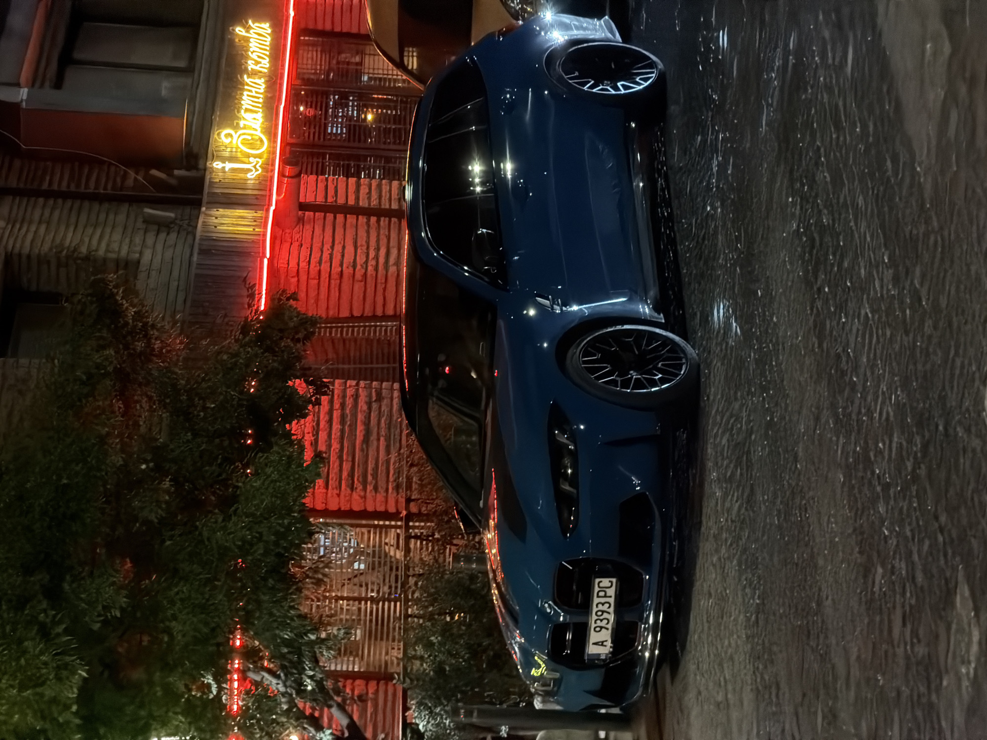 A blue BMW M3 Touring is parked on a wet cobblestone street at night in Burgas, Bulgaria. The car has a black roof and black wheels. A license plate on the front of the car reads "A 9393 PC". In the background, a building with red brick walls and neon signs is visible. One of the neon signs, written in Cyrillic, reads "Златна котвa" (Zlatna kotva), which translates to "Golden Anchor". A tree with green leaves is in the foreground to the left of the car. The streetlights and neon signs cast reflections on the wet ground.