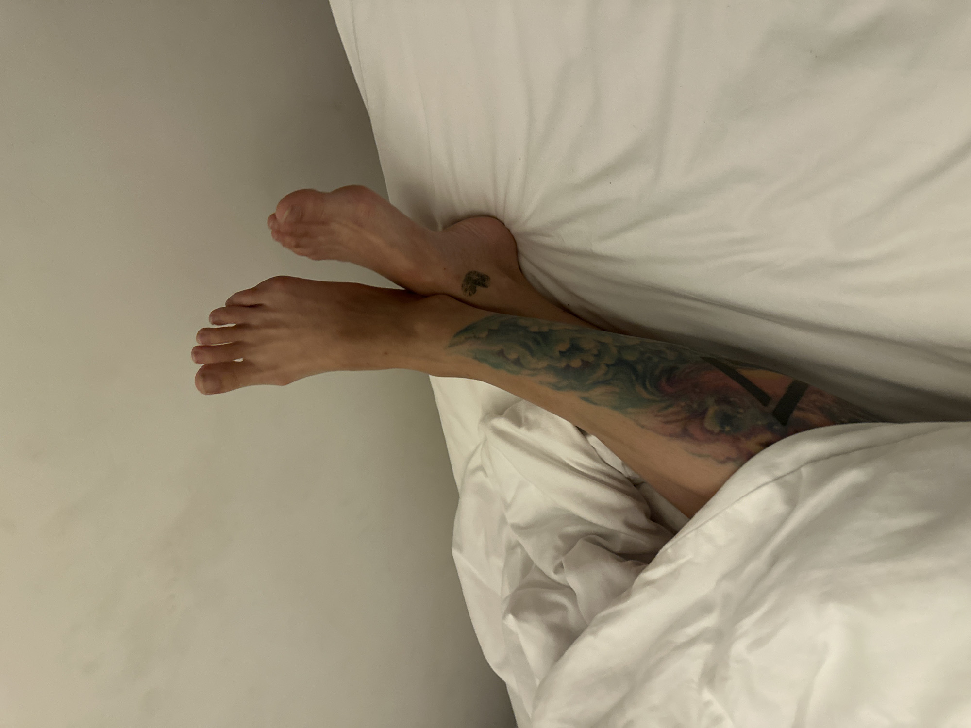 The image shows a person's legs and feet from a high-angle perspective, lying on a white bed with white sheets. The person's left leg is partially covered by the sheets, revealing a large tattoo of blue and green abstract shapes. The person's right leg is fully visible, and their foot has a small black tattoo of a heart on the ankle. The background is a plain, off-white wall. There is no indication of the location being in the city of Đà Nẵng, Vietnam.