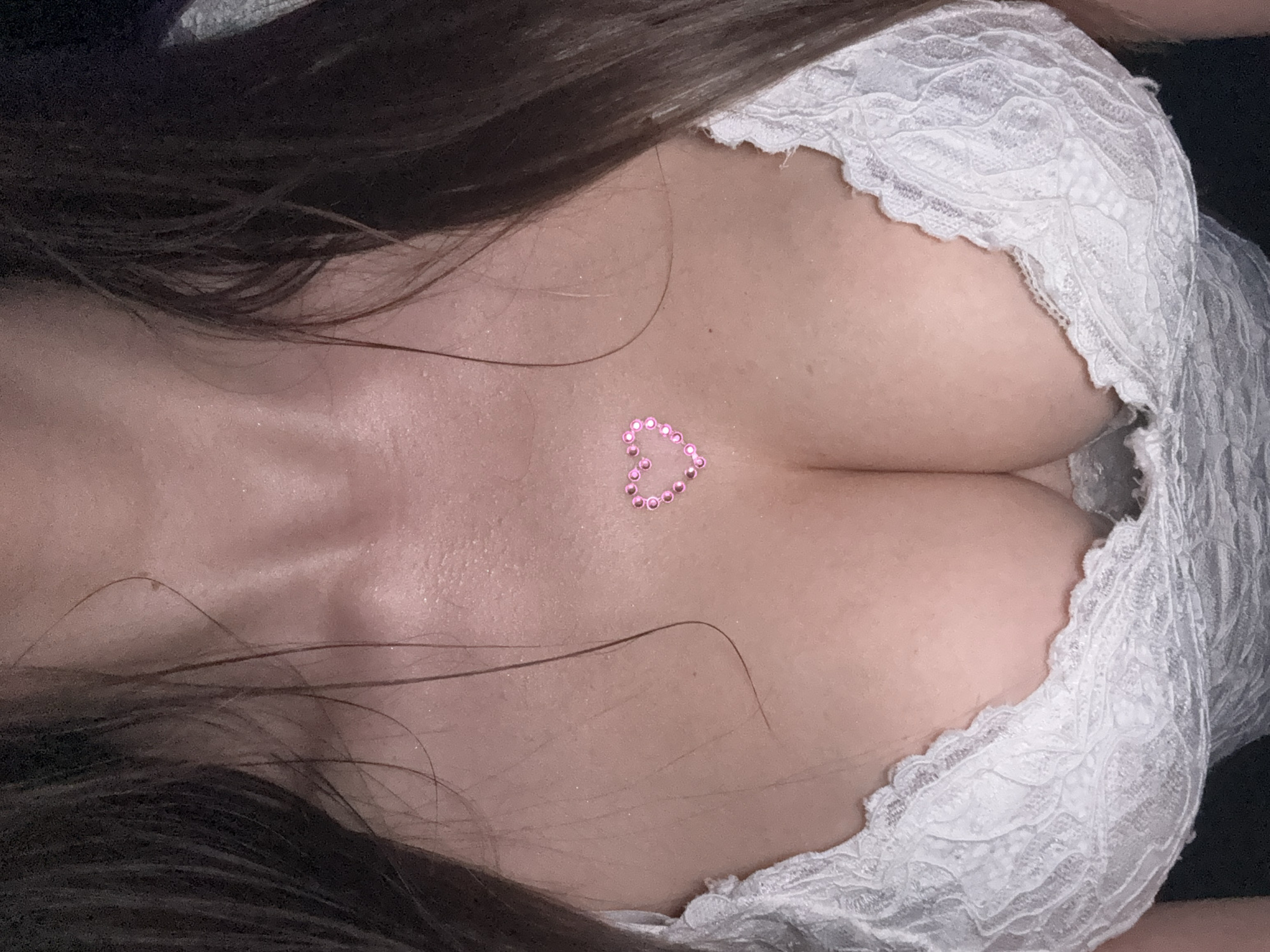 A close-up shot shows a woman's chest, cleavage visible. She is wearing a white lace garment. A small pink heart made of small rhinestones is affixed to her chest between her breasts. Her dark brown hair is visible at the top of the frame. The location is not discernible.