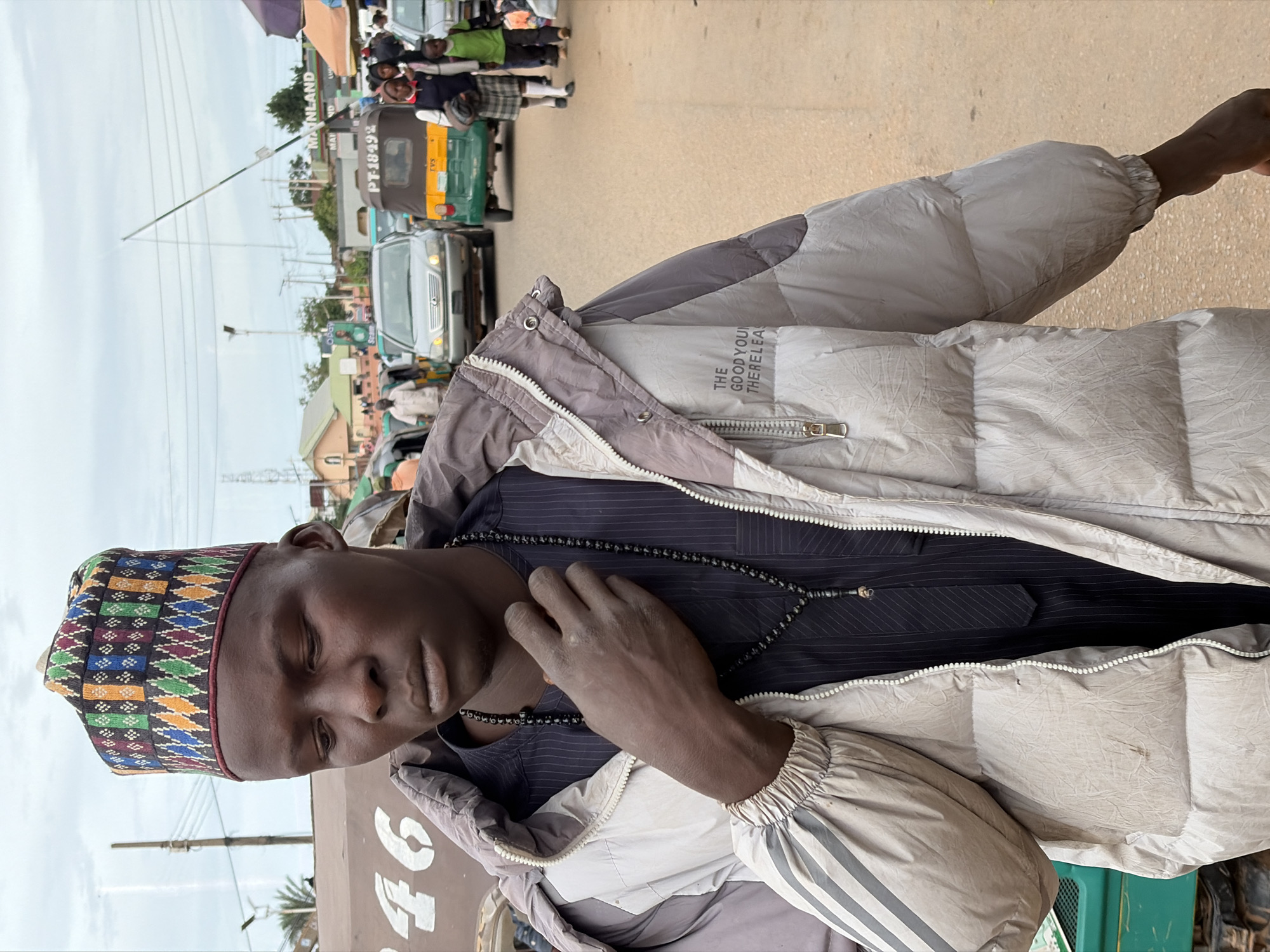 A young Black man wearing a puffer jacket and a colorful, geometric patterned cap stands in the foreground of this medium shot image. He has his left hand on his throat, and he wears a dark, pinstriped garment underneath his jacket, adorned with a long, beaded necklace. The background depicts a street scene in Jos, Nigeria, with vehicles, people, and market stalls under an overcast sky. A light-colored car with a license plate reading "PT-18492" and a green and yellow auto-rickshaw are prominent. Two children, one in a green shirt and dark pants and another in a plaid skirt and white top, walk behind the vehicles, accompanied by other pedestrians. Buildings and power lines are visible in the distance. The man's jacket has the text "THE GOOD YOUNG THERELEA" printed on the back.