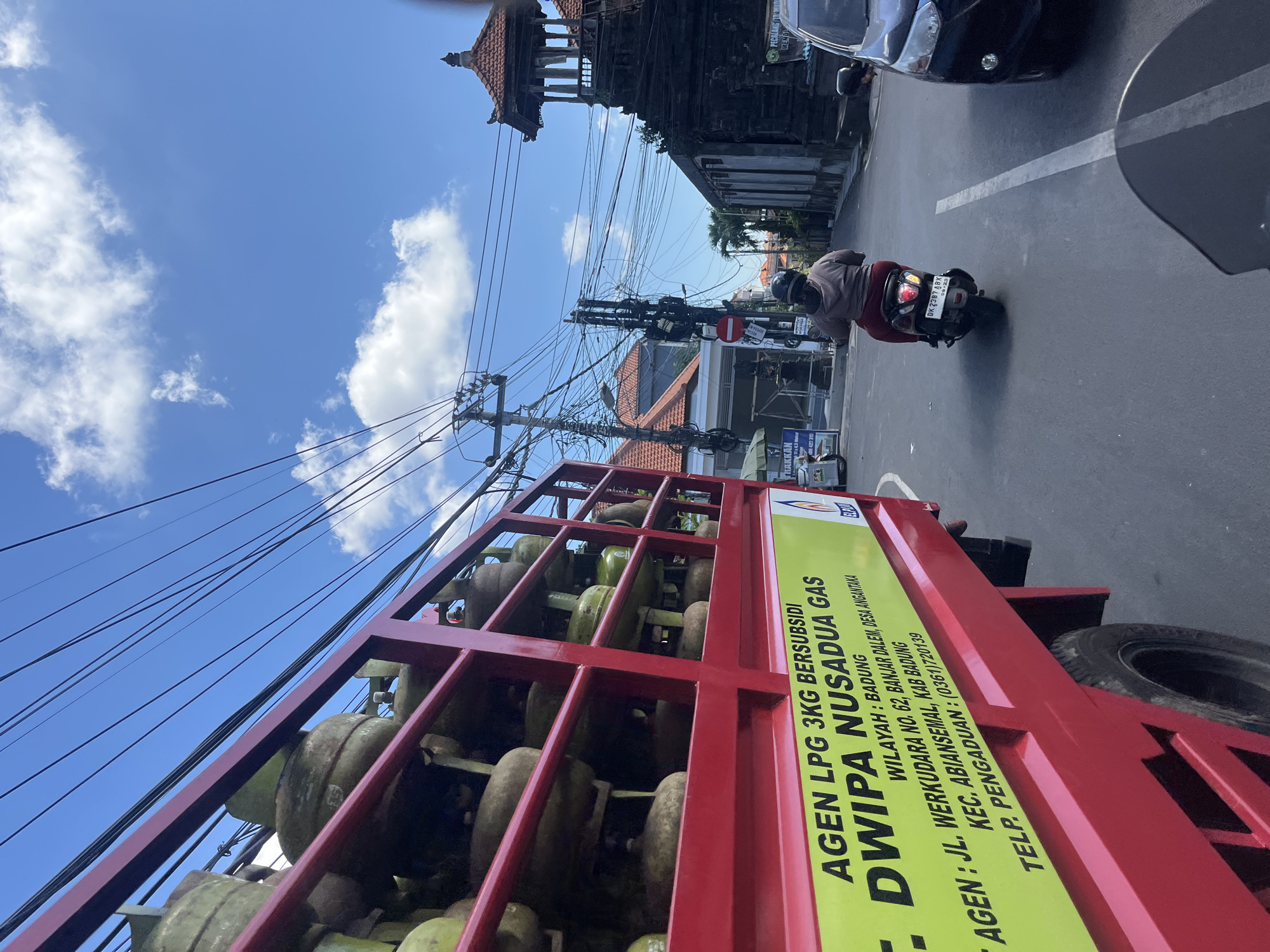 The image captures a street scene in Denpasar Barat, Indonesia, dominated by a red truck carrying numerous yellow propane gas cylinders. The truck is partially visible on the left side of the frame, with its side displaying text in yellow, including "AGEN LPG 3KG BERSUBSIDI DWIPA NUSADUA GAS", an address "JL. WERKUDARA NO. 62, BANJAR DALEM, DESA ANGKANTANA", and a telephone number "0361720139". The truck is positioned on a paved road, indicating it is likely in motion or parked.
In the middle ground, a person is riding a scooter in the same direction as the truck. The scooter's license plate is visible, reading "DK 2387 ABX". The rider is wearing a light brown hoodie and dark pants, with a helmet on. The scooter is in the lane of traffic, and a white line marks the lane division on the road.
The background reveals a street lined with buildings, some with tiled roofs. A distinctive traditional Balinese structure with a multi-tiered roof and intricate carvings is visible on the right side of the image. Overhead, a dense network of electrical wires crisscrosses the sky, typical of urban environments. The sky is blue with scattered white clouds, suggesting it is daytime. A black car is parked on the right side of the road, partially obscuring the view of the building. The overall scene depicts a busy street with transportation and commerce.
