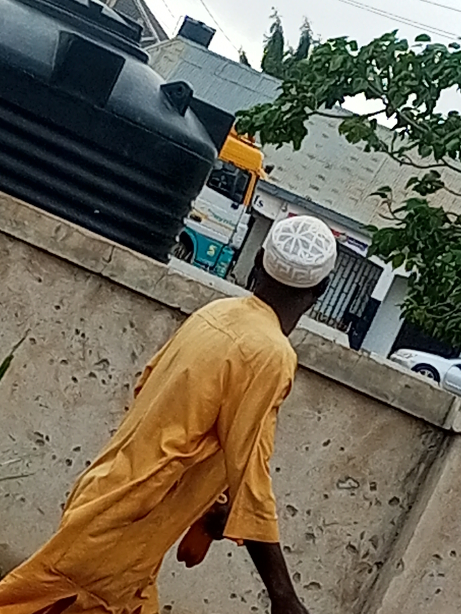 In Tumfure, Nigeria, a man wearing a yellow robe and a white kofia walks past a large black water tank. Behind him, a truck with a white and blue cab is parked near a building. The man appears to be looking to his right, towards the building, while walking. The scene is outdoors, with a gray, cloudy sky overhead and some green foliage visible in the background. The ground appears to be paved, with some rough textures and small pebbles.