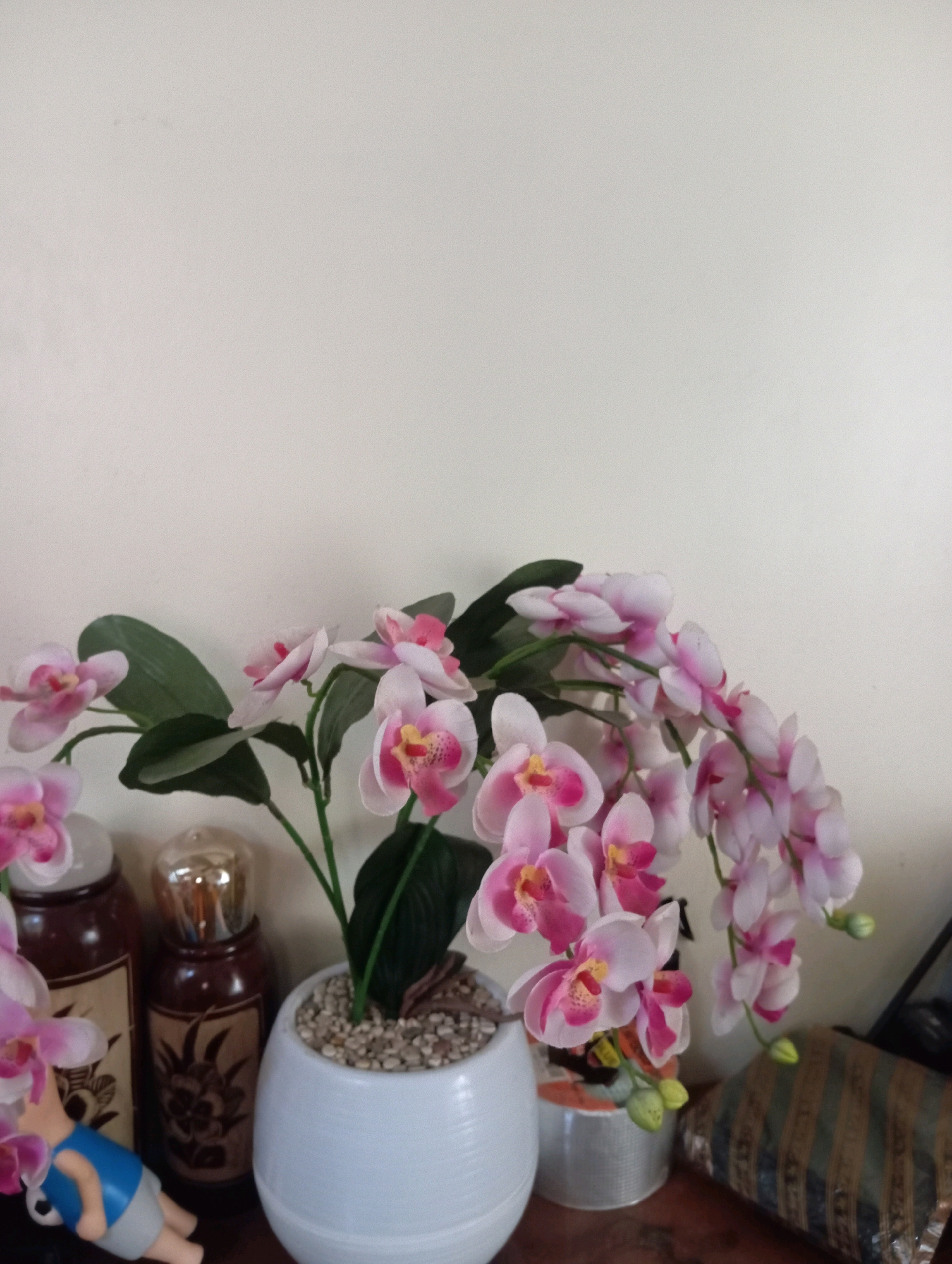 The image shows a close-up of artificial pink and white orchids in a white pot, placed on a wooden surface in front of a plain, light-colored wall. To the left of the orchid pot, there are two dark brown bottles with decorative labels and a small figurine of a person wearing a blue shirt and gray shorts. To the right of the orchid pot, there is a cylindrical object with a patterned design and a striped object lying horizontally. The lighting suggests an indoor setting. There is no visible indication of the city of Garut, Indonesia in the image.