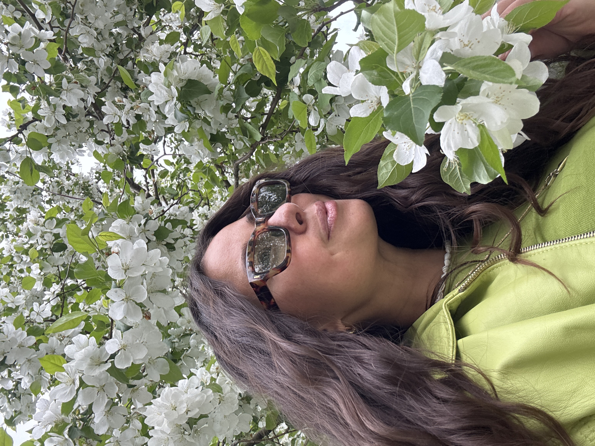 A woman with long brown hair, wearing sunglasses and a light green jacket, is looking up at a tree full of white blossoms. The tree's branches and flowers surround her face. The location is likely in Kazan, Russia.