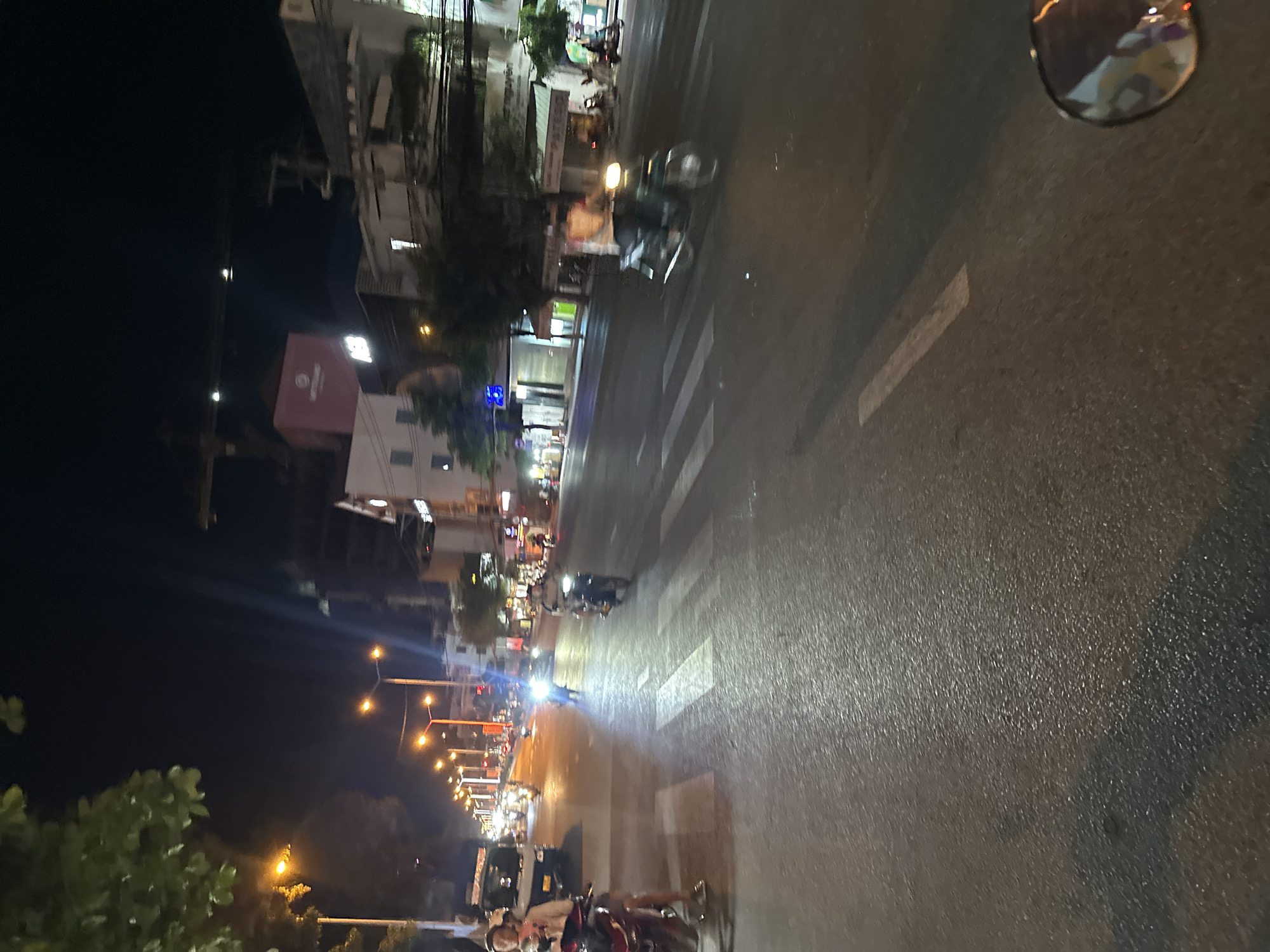The image shows a nighttime street scene in Bac Nha Trang Ward, Vietnam. A wet road surface reflects city lights and traffic. Motorbikes are the primary mode of transportation, with several visible along the street. Buildings line the road, with businesses and residential structures mixed. Streetlights illuminate the scene, and a pedestrian crossing is marked on the road. A vehicle mirror is visible on the right side.