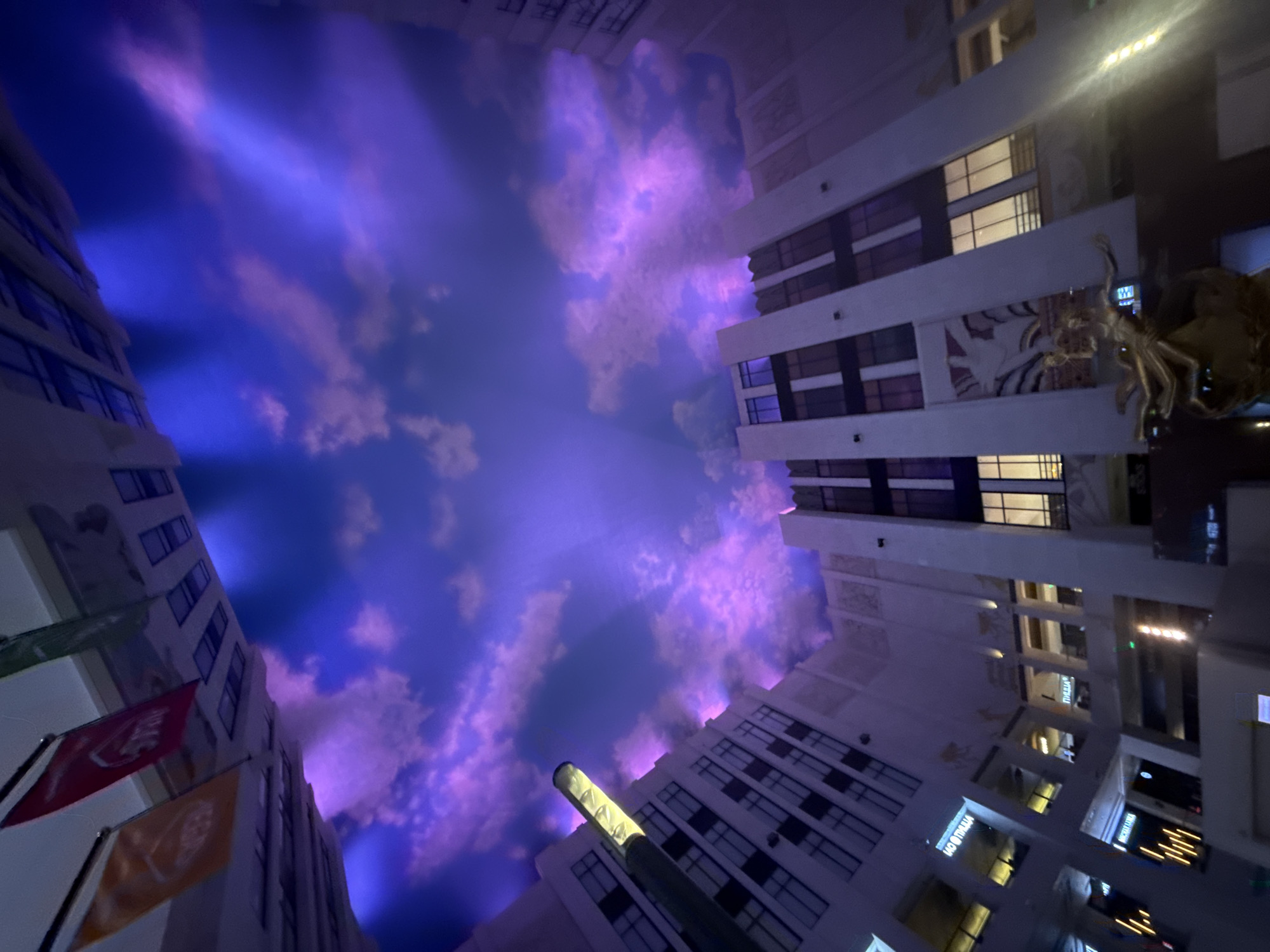 The image shows a view looking upwards towards a ceiling designed to resemble a purple and blue sky with clouds. Surrounding the open space are the exteriors of several multi-story buildings, designed in a somewhat art deco style with geometric patterns and vertical lines. Several flags are visible hanging from the upper edges of some buildings. A tall, cylindrical light fixture extends upwards from one of the buildings. Further down, there are windows, illuminated interiors, and some decorative elements on the building facades, including what appears to be a statue of a golden figure in the foreground. The overall impression is of an indoor atrium or public space with an artificial sky. The location is identified as Khimki, Russia.