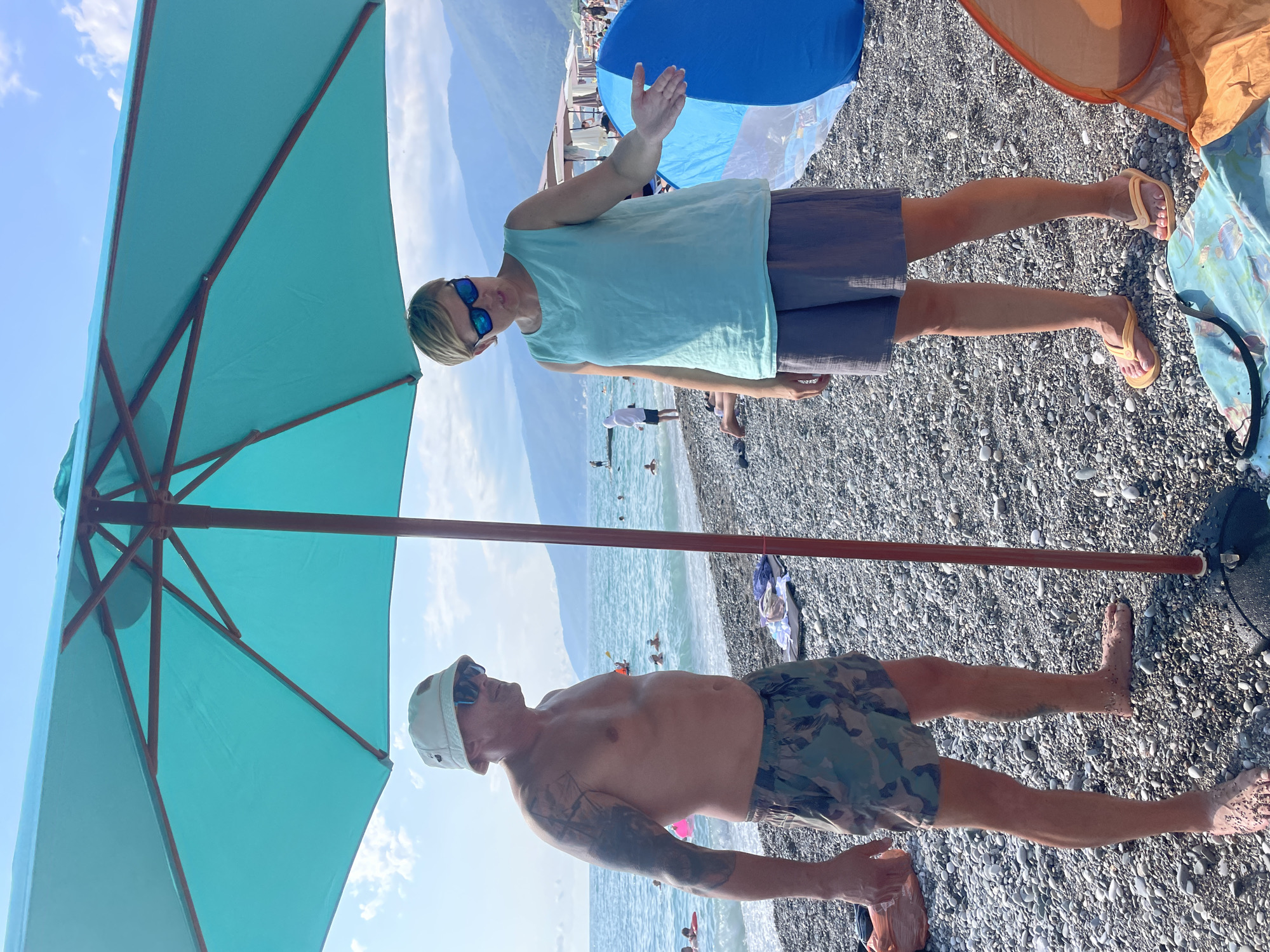 This media file captures two individuals on a pebbly beach in Gagra, Abkhazia. A large turquoise umbrella shades one person, a man wearing a light-colored bucket hat, sunglasses, and camouflage swim trunks. He has tattoos on his left arm. The other individual, standing under a blue and orange umbrella, is a person wearing sunglasses and a light blue top with dark shorts. Their arms are raised as if waving or gesturing. The sea is visible in the background, with several people in the water. The sky is bright blue with some clouds, suggesting it is daytime with good weather. The overall scene conveys a relaxed beach atmosphere, likely during a vacation or outing. There is no visible text in the media file.