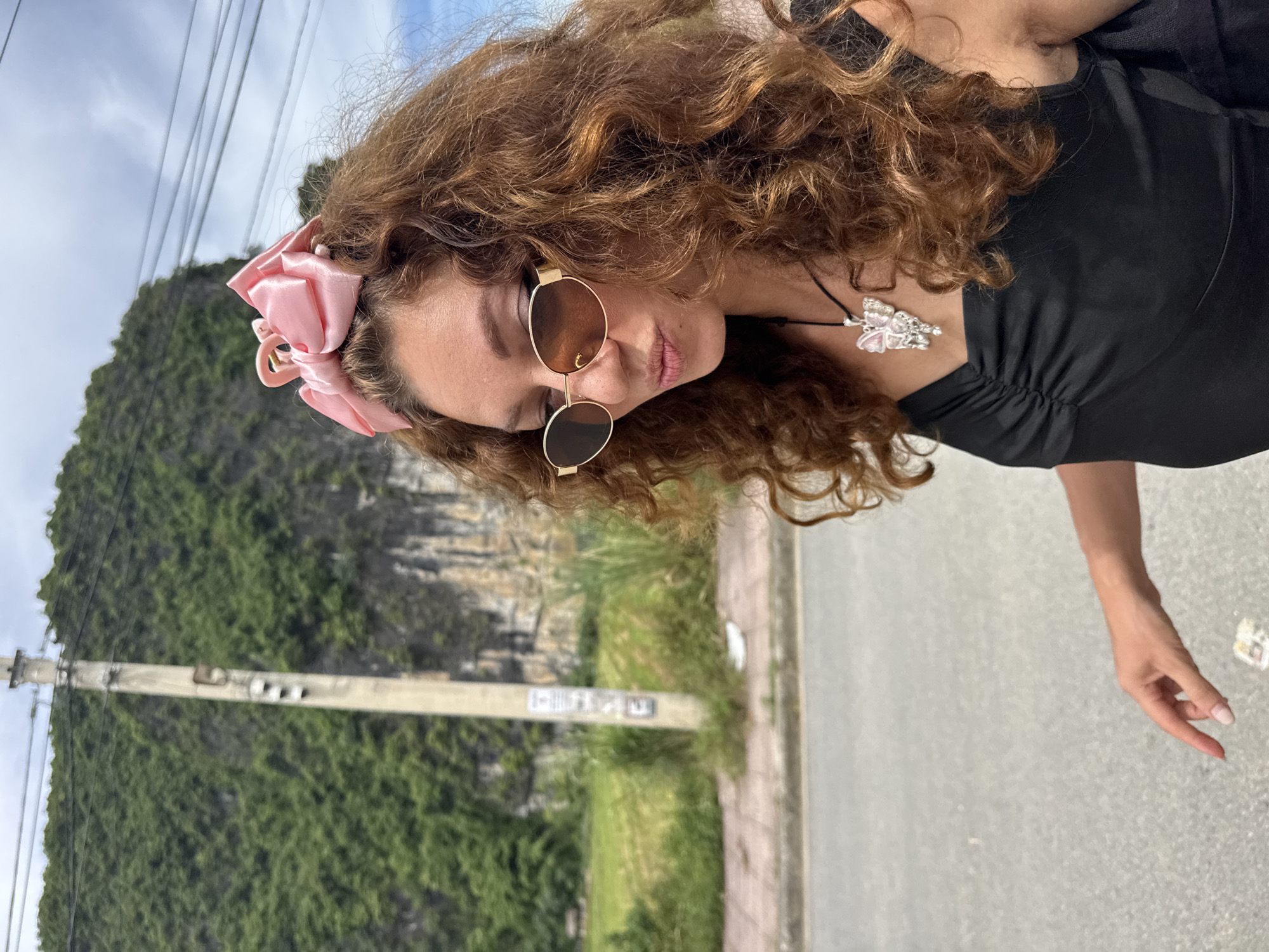 A young woman with curly brown hair is pictured in Nam Hoa Lư Ward, Vietnam. She is wearing a black dress, sunglasses, and a pink bow in her hair. She is also wearing a necklace with a butterfly pendant. The woman is standing in front of a telephone pole and a lush green mountain. The background also shows a sidewalk and a road. The sky is cloudy.