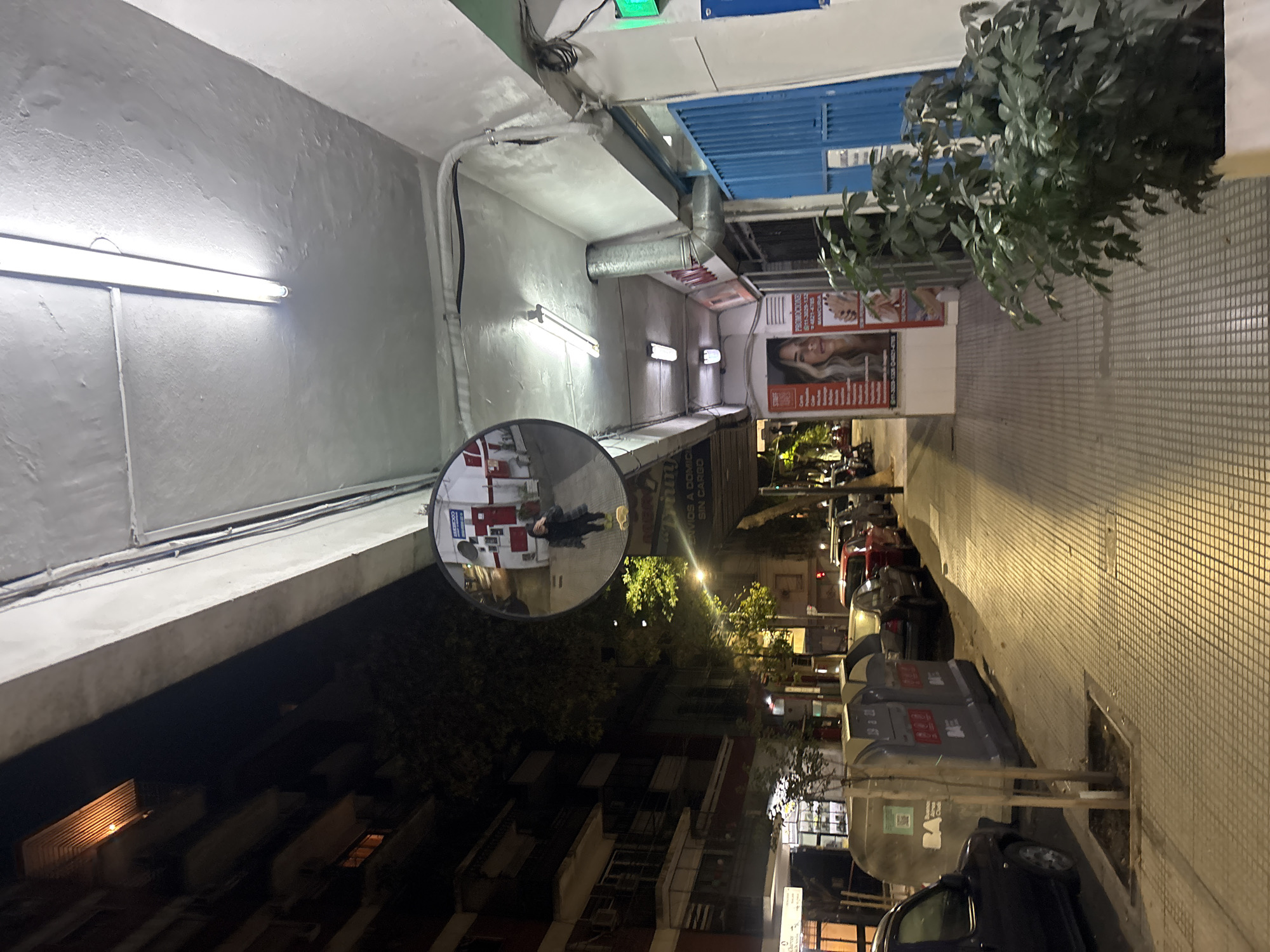 The image depicts a narrow street in Buenos Aires, Argentina, at night. On the right side of the frame, a tiled sidewalk runs along the street. Two gray BA branded recycling bins stand near the curb, next to a dark-colored car. Across the street, a series of buildings are visible, including one with illuminated windows. A convex mirror mounted on the side of a building offers a distorted reflection of the street behind the viewer. The mirror captures a figure standing on the sidewalk, in front of a business. The street is lit by overhead fluorescent lights that run along the ceiling of the building overhang. A tree is seen on the right of the image. A storefront has a sign advertising nail services. The location appears to be a commercial area with both residential and business structures.