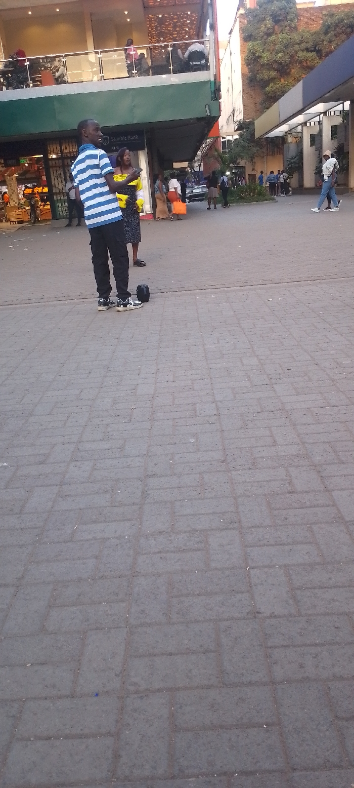 The image captures an outdoor urban scene, likely a public square or pedestrian area in front of commercial buildings. The ground is paved with rectangular grey and dark grey interlocking bricks, showing some signs of wear and dirt.
In the foreground, a man stands facing slightly to the right, looking forward with a slight smile or smirk. He is wearing a blue and white horizontal striped short-sleeved shirt, black cargo pants, and black and white sneakers. To his right, near his feet, is a black object that resembles a single wheel of a hoverboard or a unicycle-like personal transporter.
Behind the man, a crowd of people is visible. To the man's left and slightly behind him, a woman with long dark hair, wearing a yellow top and a dark patterned skirt, is looking towards the left, holding a phone to her ear or hand. Further into the midground, several other individuals and small groups of people are standing or walking. Some are carrying bags; one person on the right side of the midground carries an orange bag. In the background, what appears to be a street with parked vehicles can be seen, and more pedestrians are present.
The buildings on the left side of the image are multi-story commercial structures. The lower level has large windows, and signage for "Stanbic Bank" is clearly visible. Above this, there's a green canopy or awning, and a second story features an open balcony or terrace with glass railings, where more people are visible, some seated. To the right of these buildings, there's a denser urban environment with trees and other structures. The lighting suggests it is daytime, possibly late afternoon, given the warmth of the light. The overall atmosphere appears casual and busy, typical of a commercial district or public gathering space.