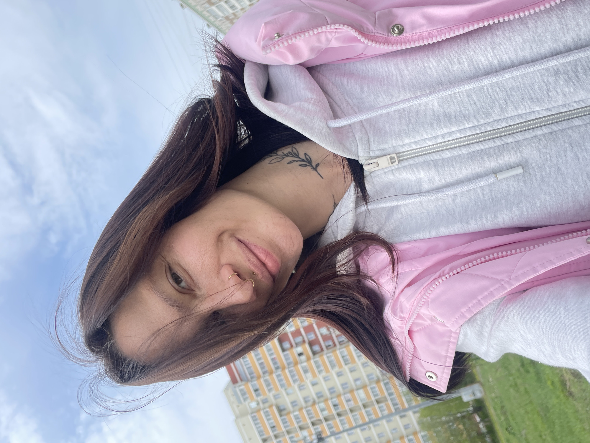 A selfie of a woman with brown hair, a gold nose ring, and a leaf tattoo on her neck is in the foreground. She is wearing a grey hooded sweatshirt and a pink quilted vest. Behind her is a multi-story apartment building with a beige and red facade. A patch of green grass and a cloudy sky are visible. The location is consistent with a residential area in Izhevsk, Russia.