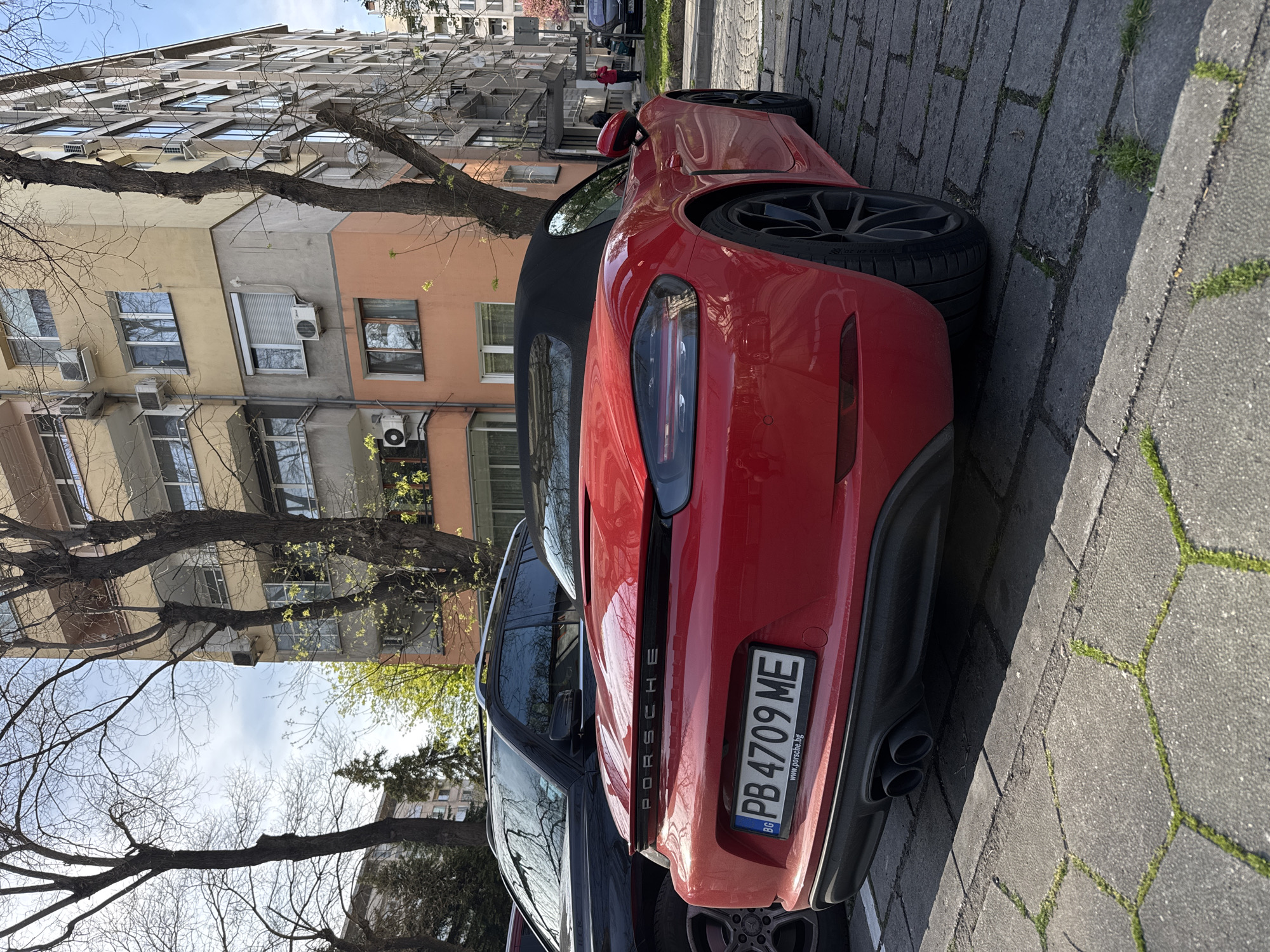 A red Porsche sports car is parked on a gray paved street alongside a dark-colored SUV. The license plate on the Porsche reads "PB 4709 ME." In the background, there is a multi-story building with various colored facades and numerous windows, likely an apartment complex. Bare tree branches are visible in the foreground and partially obscure the building. The photo appears to have been taken during the daytime. Based on the Bulgarian license plate and the general architecture, the location is likely somewhere in Plovdiv, Bulgaria.