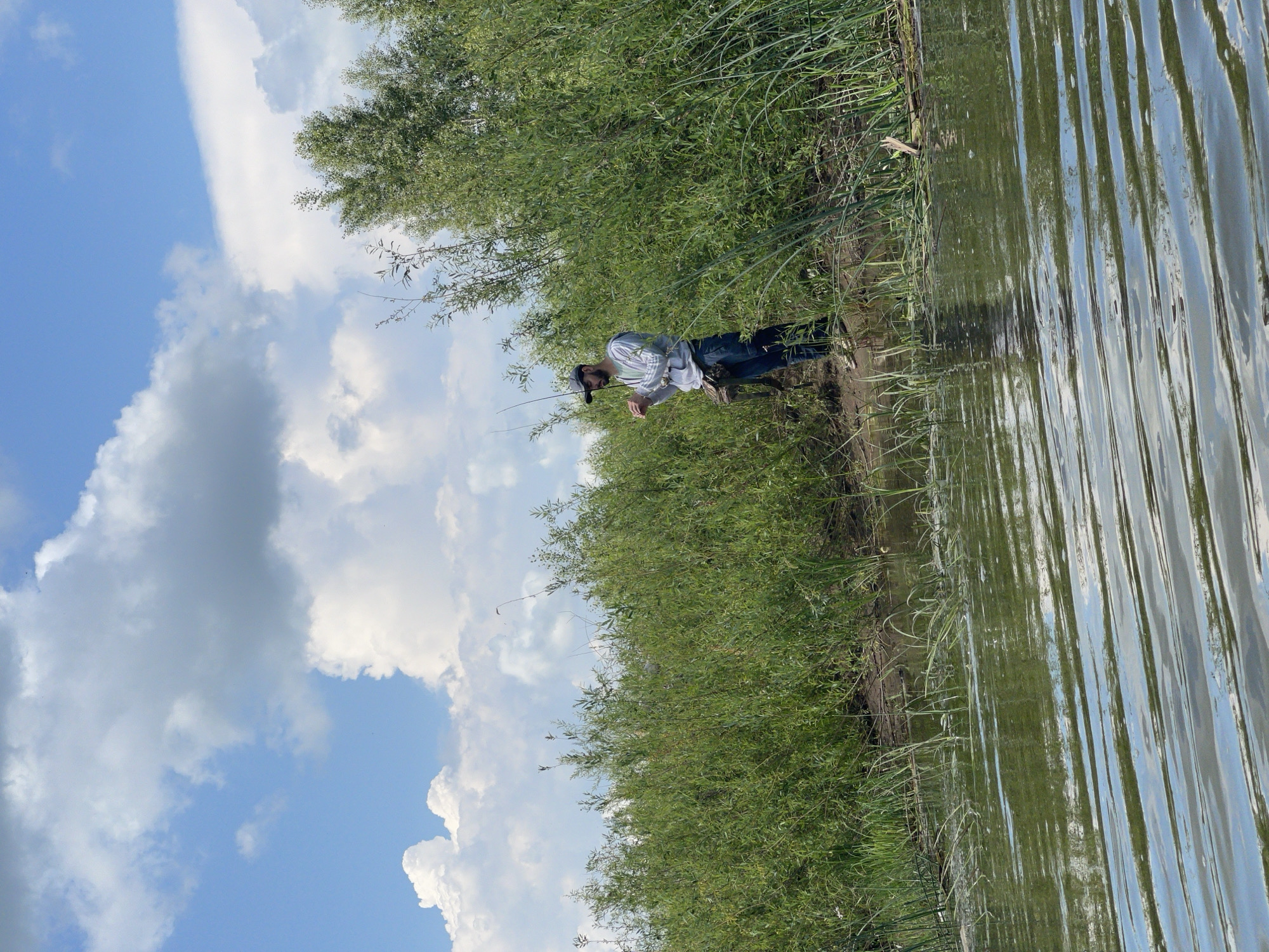 A man stands on the muddy bank of a body of water, holding a fishing rod. He is wearing a gray hooded sweatshirt, blue jeans, a dark baseball cap, and green rubber boots. A dark fishing vest is slung across his chest. Behind him, dense green foliage lines the bank, and a line of trees extends into the background. The sky above is blue with scattered white clouds. The foreground shows the rippling surface of the water, reflecting the sky and surrounding vegetation. The location is identified as being in the city of Nezhinka, Russia.
