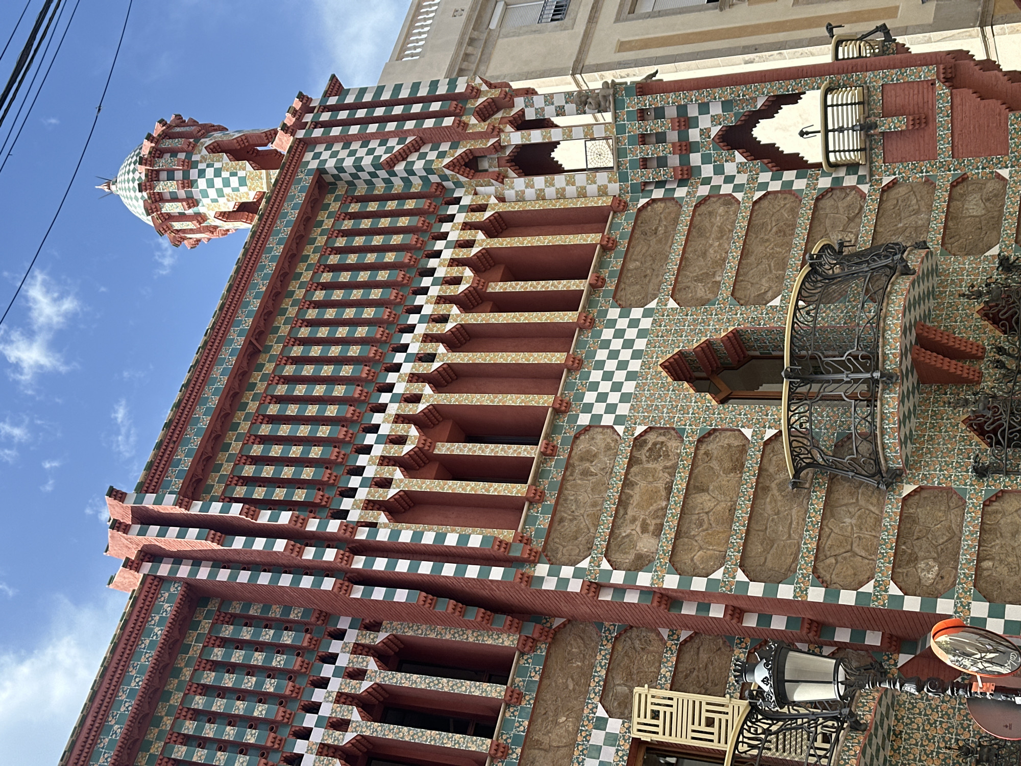 The image shows a detail of the exterior of Casa Vicens, located in Barcelona, Spain. The building's facade is highly decorated with colorful ceramic tiles and brickwork, featuring a checkerboard pattern of green and white, and red brick accents. The architecture includes arched windows, balconies with ornate metal railings, and a distinctive tower with a tiled dome. A street lamp with a glass lantern is visible in the foreground, along with a traffic mirror. The sky above is blue with scattered clouds. No people are visible in the image.