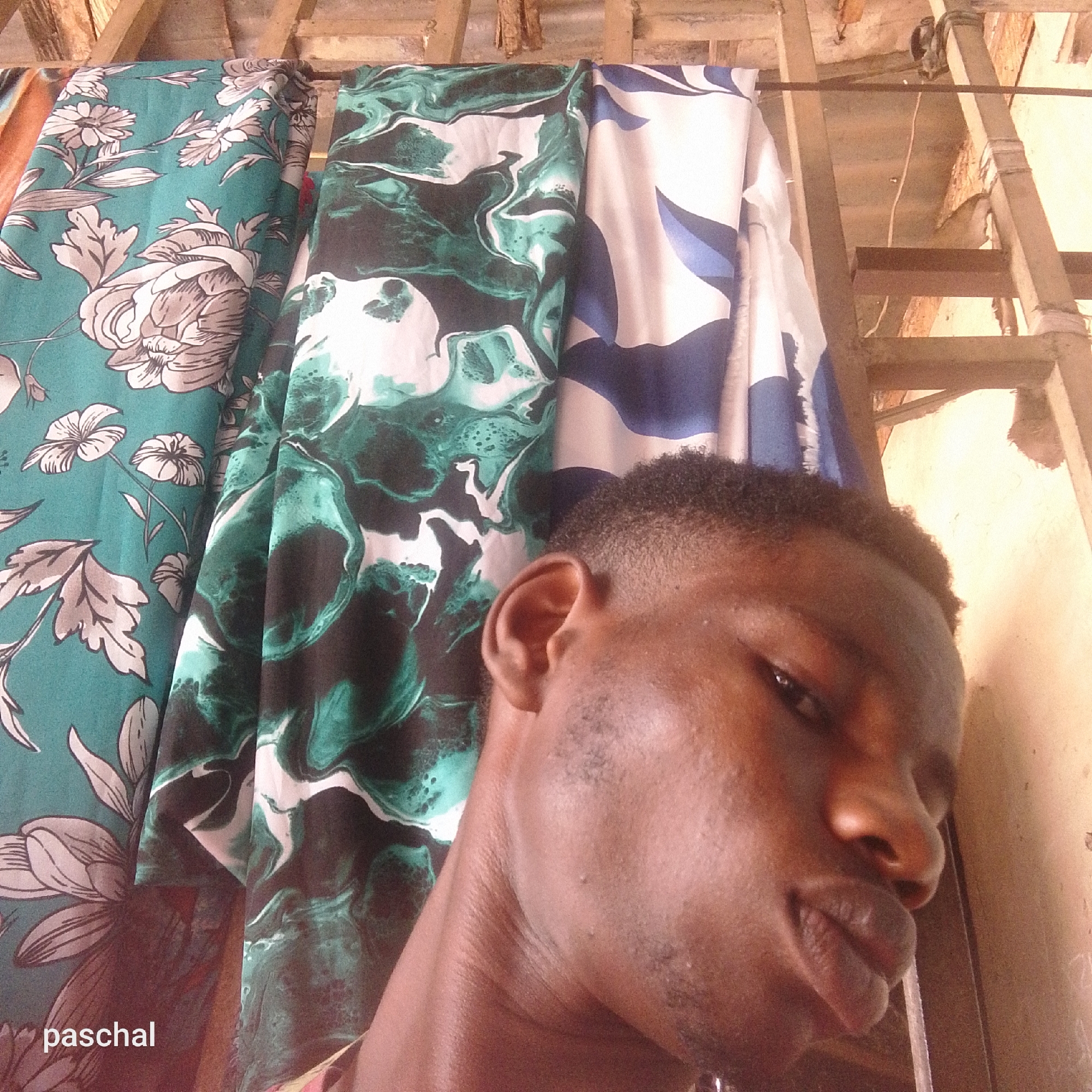 The image is a close-up selfie of a dark-skinned person, likely male, with short, dark hair. The subject's face is partially illuminated, showing details like stubble. The background consists of draped fabrics with varying patterns: one with large, stylized floral designs in teal and grey, another with a swirling marble pattern in shades of green, and a third with a white and blue abstract design. A wooden structure, possibly a ladder or scaffolding, is visible in the upper right corner. "paschal" is written in the lower-left corner, likely a watermark. The location is stated to be in the city of Nsugpe, Nigeria. No specific details within the image confirm this, however. No discernible actions or interactions are occurring in the image.