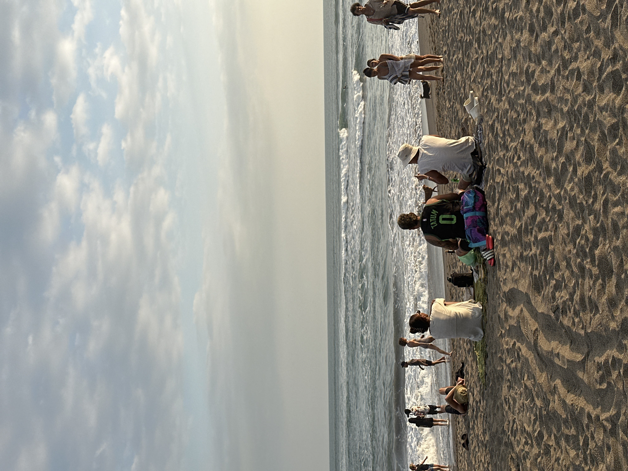 In North Kuta, Indonesia, a sandy beach is depicted with the ocean waves rolling in under a partly cloudy sky. Several people are scattered across the beach. In the foreground, three individuals are seated on the sand, facing the ocean. One wears a green basketball jersey with the number "0" and "TATUM" visible on the back, another a white bucket hat, and the third a white t-shirt. A small dog is near the seated individuals. Further back, closer to the water's edge, a group of people are standing and walking. Some are in the shallow water. The sun appears to be low in the sky, casting shadows on the sand.