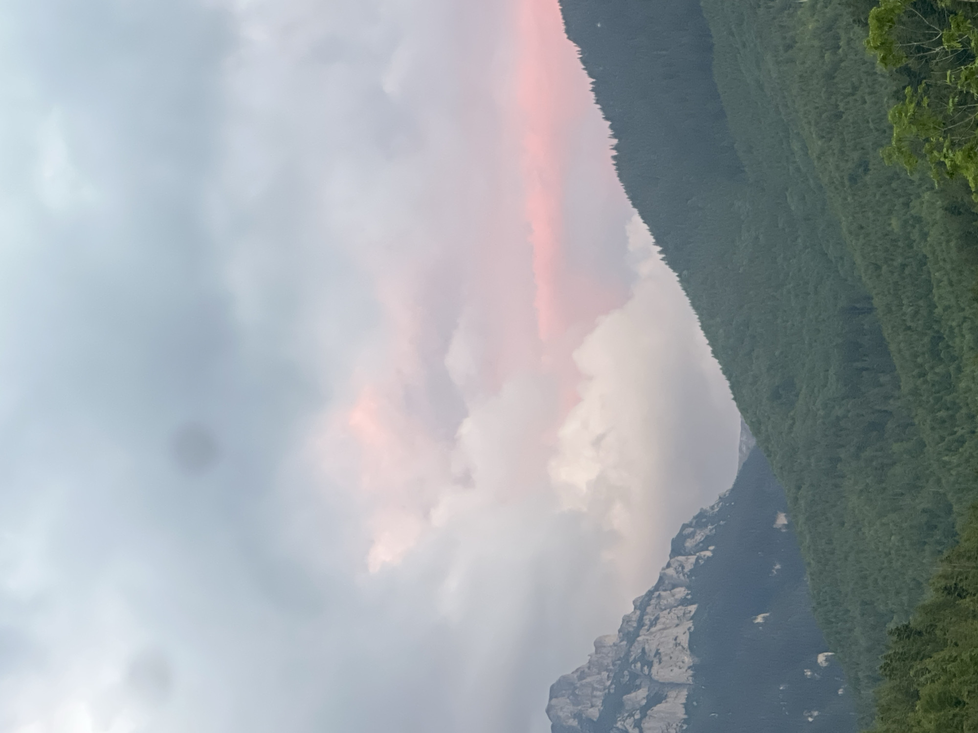 The image shows a panoramic view of a mountainous landscape under a cloudy sky. In the foreground, lush green forests cover the slopes of rolling hills. The middle ground is dominated by a jagged, rocky mountain peak on the left, with dark green coniferous trees growing on its lower slopes. To the right of this peak, another, more rounded, tree-covered mountain rises, its silhouette sharp against the sky. The sky itself is filled with dramatic clouds, a mixture of grey and white, with a soft band of pinkish light visible in the upper portion, suggesting either sunrise or sunset. There are no people or man-made structures visible in the image. The overall impression is one of a remote and natural wilderness.
