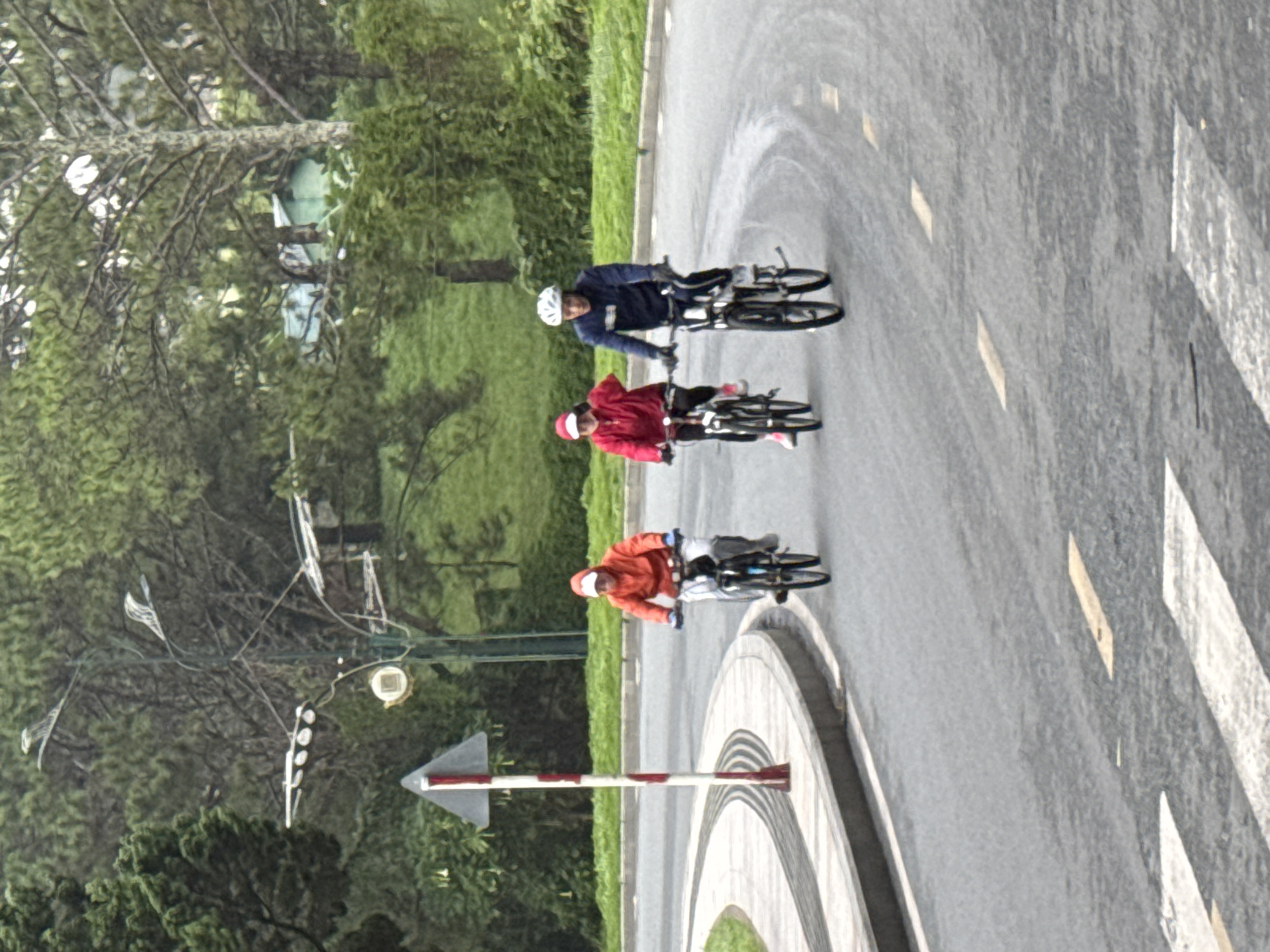 Three individuals are cycling on a paved road in Phường Xuân Hương, Đà Lạt, Vietnam. The cyclist on the left wears an orange hooded jacket and white pants, riding a black bicycle. In the middle, a person in a red hooded jacket and dark pants, with a white cap, is on a dark bicycle. The cyclist on the right wears a dark blue jacket and pants, with a white helmet, riding a dark bicycle. The road curves to the left, bordered by a concrete curb and a grassy slope with trees in the background. A traffic signpost with red and white stripes is positioned to the left of the road. The overall environment appears to be a park or a scenic route.