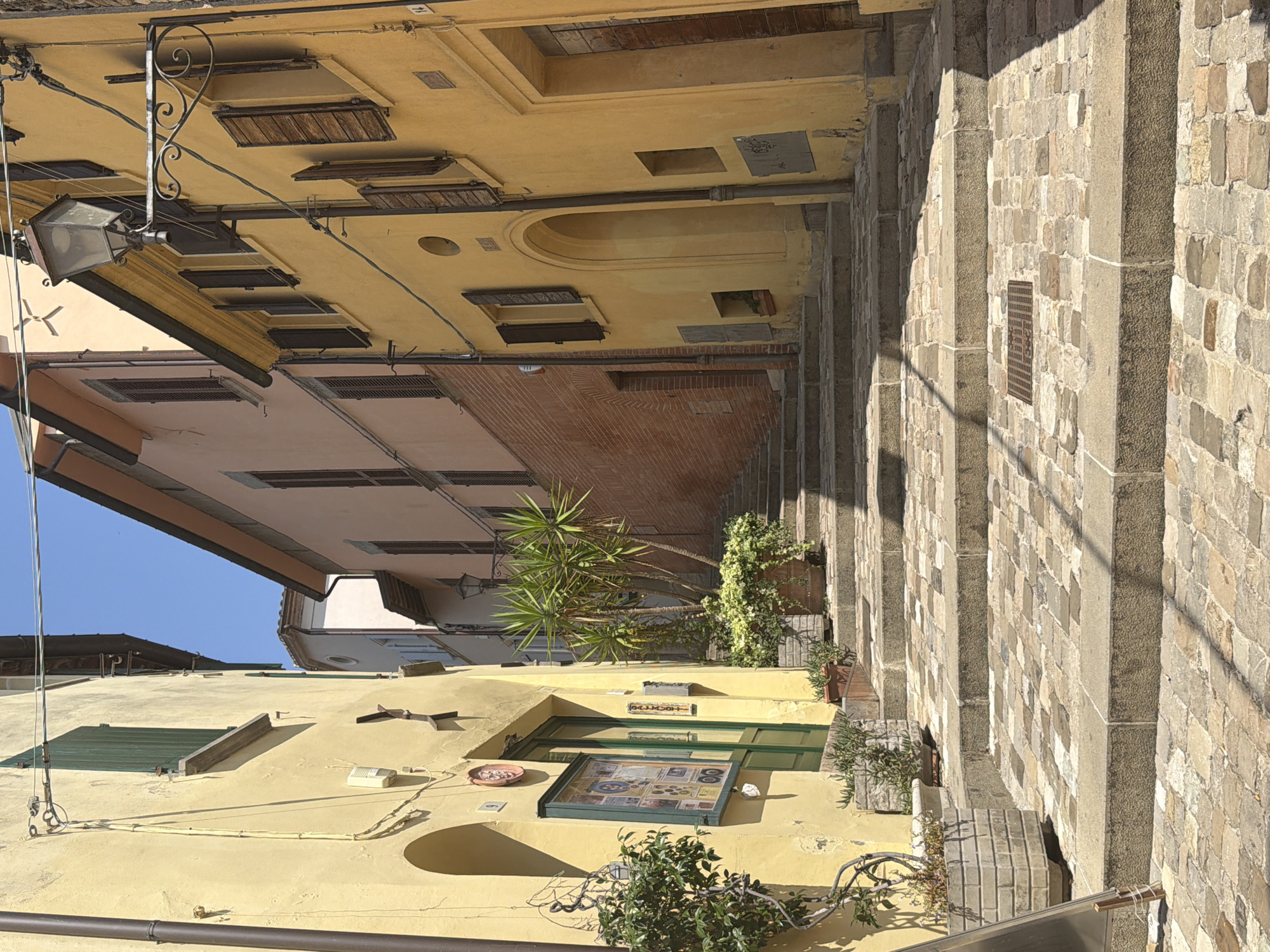 The image shows a narrow, cobblestone street in the historic center of Santarcangelo di Romagna, Italy. The street ascends a steep incline, forming a staircase lined with old buildings. The buildings on either side are painted in warm, earthy tones of yellow and pale pink, with traditional wooden shutters on the windows. The architecture suggests a Mediterranean setting, likely dating back several centuries.
On the left side of the street, a yellow building has a green door and a display case mounted on the wall. A potted plant sits near the entrance. The building on the right also features wooden shutters and a wrought-iron lamppost extending over the street. A large potted plant with spiky leaves is visible on the steps, adding a touch of greenery to the scene. The cobblestone pavement is a mix of light and dark stones, worn smooth by time. The sky is clear and blue, indicating a sunny day. No people are present in the frame, creating a sense of stillness and timelessness. The overall impression is one of quaint, old-world charm.