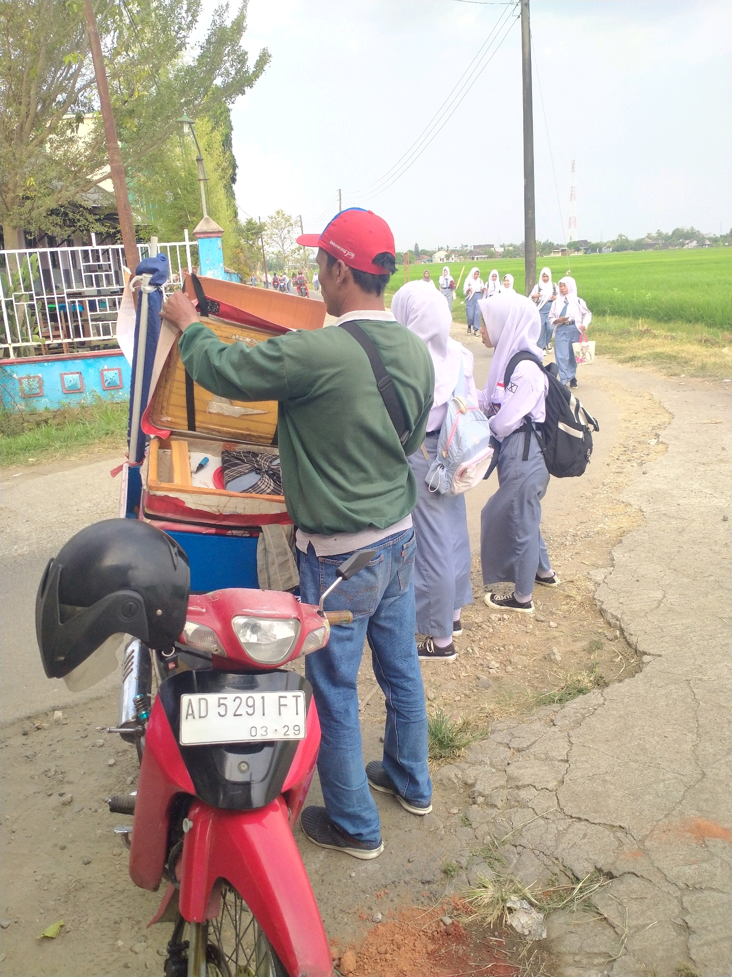 In the city of Begajah, Indonesia, a man wearing a red cap and green long-sleeved shirt stands beside a red motorcycle. The motorcycle has a black helmet attached to its front. The man is interacting with the contents of a wooden cart attached to the motorcycle. In the background, a group of young women in white hijabs and school uniforms walk along a dirt road bordered by a vibrant green rice field. The environment appears rural, with a few houses and trees visible in the distance. The sky is bright and overcast.