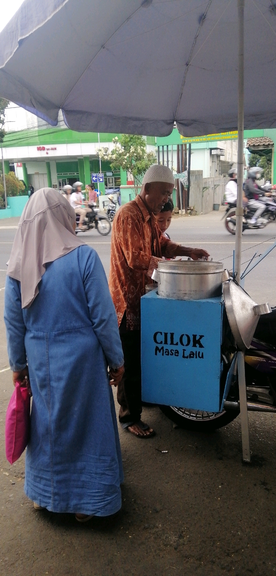 The image captures a street scene in Ajibarang Wetan, Indonesia, likely near a healthcare facility indicated by signage that includes "IGD 24 JAM," translating to "Emergency Room 24 Hours." A food vendor, an elderly man wearing a white peci cap and an orange patterned shirt, is seen attending to his cart. Beside him, a young boy, possibly his grandson, looks on. The cart, painted blue, displays the text "CILOK Masa Lalu," identifying the food being sold as "Cilok Past." A woman wearing a blue abaya and a beige headscarf stands to the left, holding a pink bag, and appears to be a customer. In the background, traffic is visible, with several motorcycles and riders on the road, suggesting a typical urban environment. The overall atmosphere is that of a casual transaction on a roadside, under a large gray umbrella that provides shade.