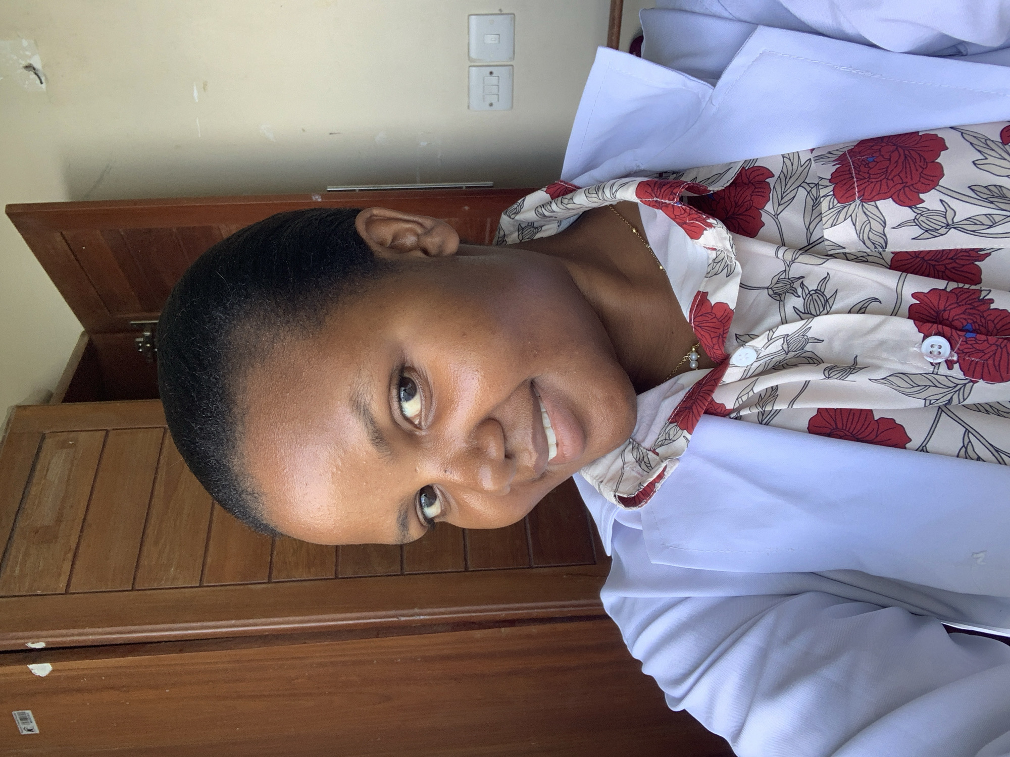 A woman is photographed indoors. She is wearing a white lab coat over a floral-patterned blouse. The interior includes a wooden cabinet behind the woman and light switches on the wall. While not explicitly visible, the location is presumed to be in Dar es-Salaam, Tanzania, as requested in the prompt.