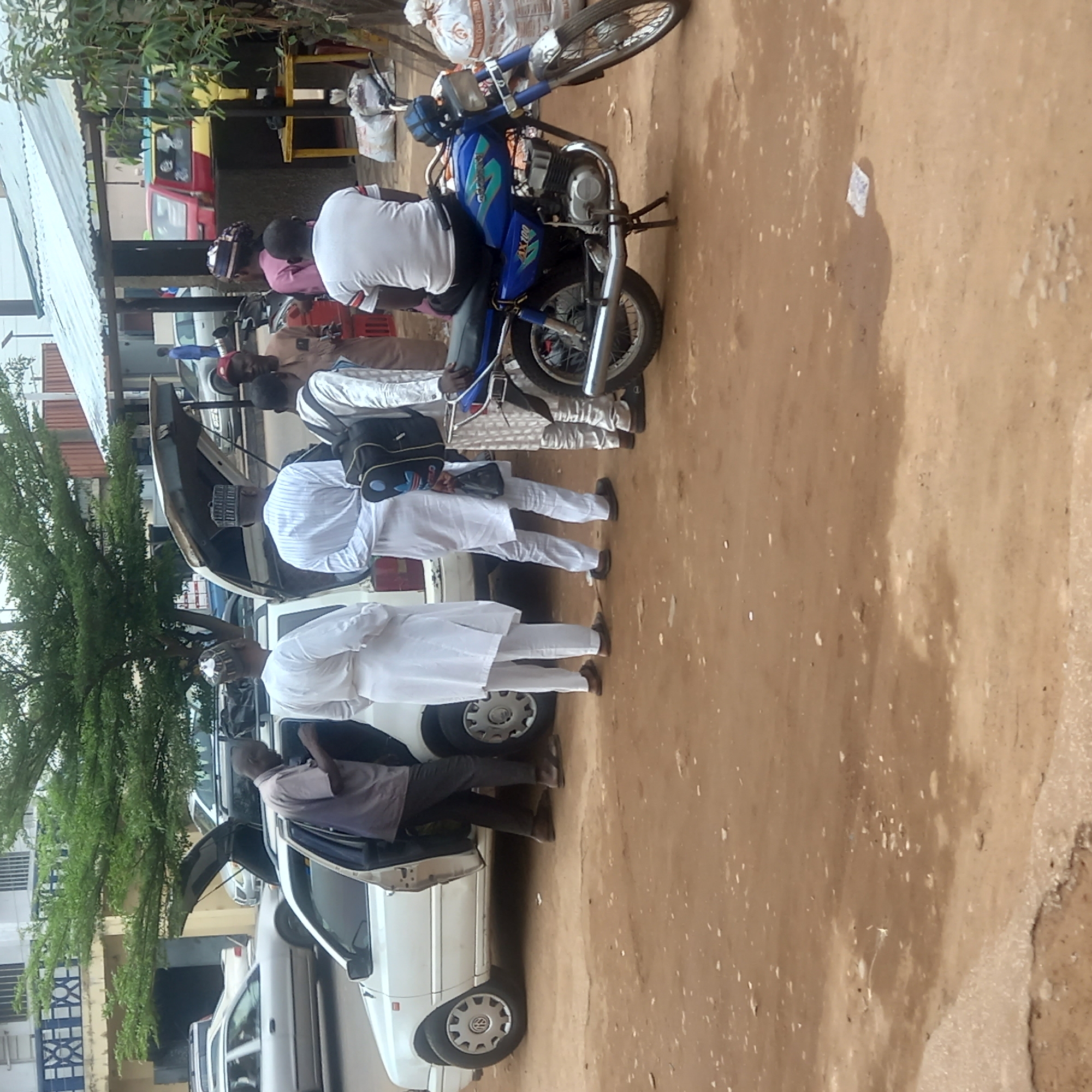 The scene takes place in Dadi Riba, Nigeria. Several men, some wearing white traditional attire, are standing near a row of white cars with open doors and trunks. One man sits on a blue motorcycle. The ground is dirt. Buildings are visible in the background.