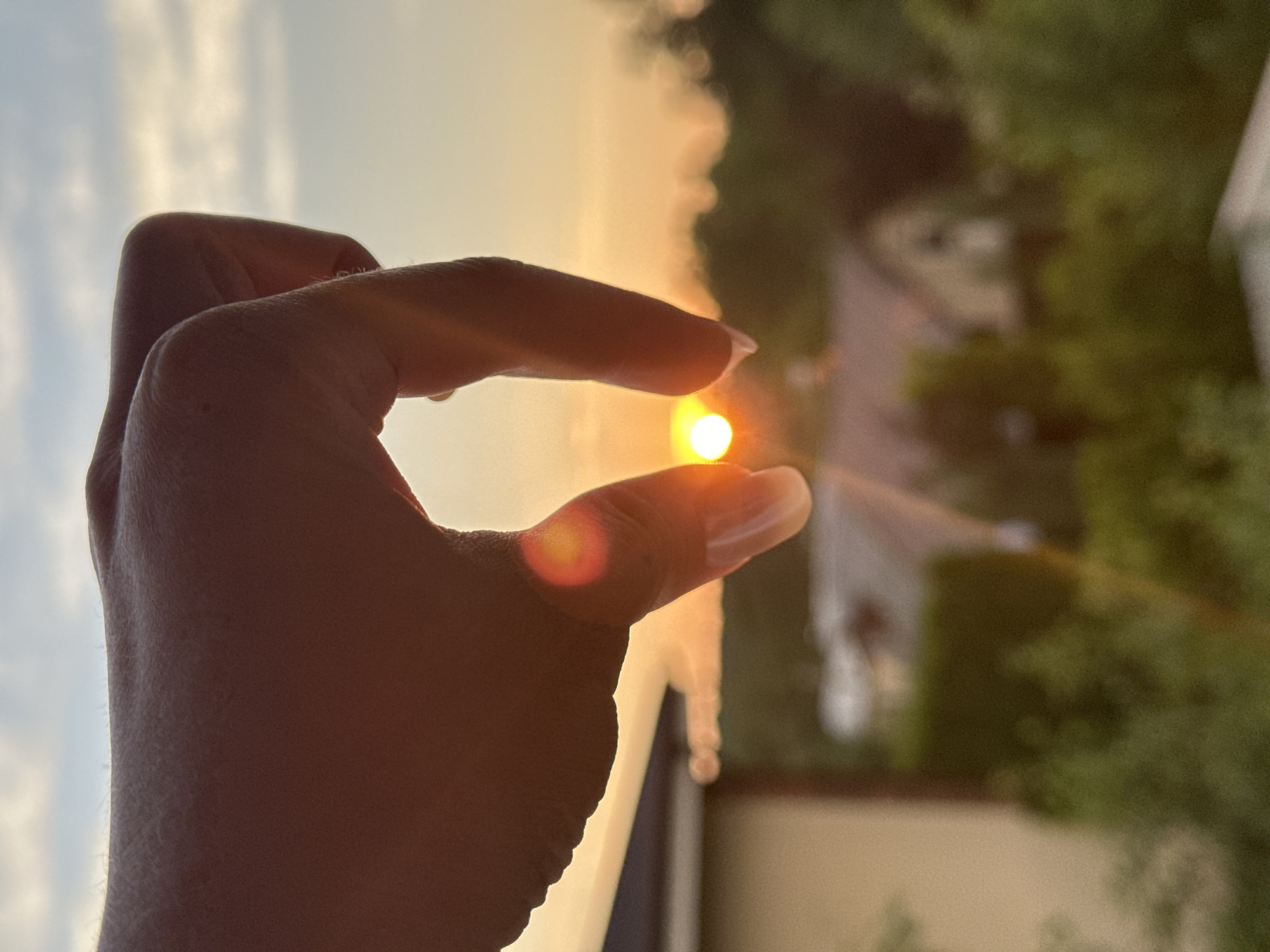 A close-up shot captures a human hand, silhouetted against a sunset. The sun, a bright orange disc, is held between the thumb and index finger of the hand, appearing as if it is being grasped. The background is a blur of warm-toned sky, soft green foliage, and the indistinct shapes of buildings, suggesting a suburban or semi-rural setting. The image's composition creates a symbolic or artistic impression of holding or containing the sunset. There is no identifiable information about the location in Nizhny Novgorod, Russia.