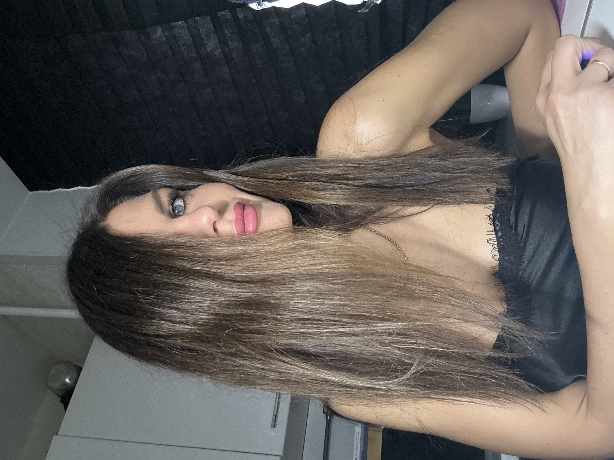 A woman with long brown hair and green eyes is posing for a picture. She is wearing a black top with lace. The background is a combination of black curtains and a white wall with a kitchen area. The location is Chaikovsky, Russia.