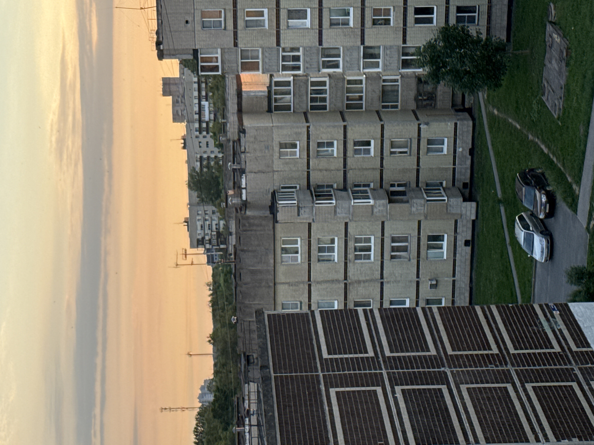 An aerial, eye-level view captures the evening sky over Kirishi, Russia, casting a warm, orange glow on apartment buildings and surrounding vegetation. The sky is partially obscured by streaks of pale clouds, suggesting the sun is near the horizon. In the foreground, a large, multi-story apartment building with dark, textured panels featuring a white geometric pattern dominates the left side of the frame. To its right, another, lighter-colored apartment building with a grid-like facade is visible, its windows appearing dark. Further in the background, a line of similar apartment buildings stretches across the landscape, interspersed with trees and topped with television antennas and radio towers. The ground level reveals a patch of green grass, a paved road, and two parked cars: a silver sedan and a dark SUV. A lone tree stands in the grassy area. The scene depicts a typical residential district at dusk.