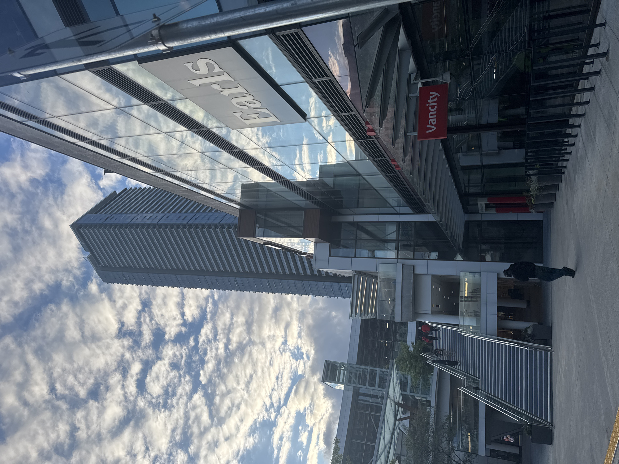 The image shows a daytime exterior scene in Burnaby, Canada, likely in a commercial or urban development area. Dominating the background is a very tall, modern high-rise building with a repetitive pattern of balconies or window sections, suggesting it is a residential or office tower. In the foreground and mid-ground, a contemporary multi-level building features a glass facade that reflects the cloudy sky. A prominent sign on this building reads "Earls" in large white lettering. Below the Earls sign, another red sign reads "Vancity". A wide staircase with metal railings ascends to the left of the frame, with several people visible on it. A lone individual walks away from the camera on the paved ground in the foreground, to the right of the staircase. Planters with greenery are placed along the pathway. The environment appears clean and well-maintained, characteristic of a developed urban setting. The sky is partly cloudy with scattered white clouds against a blue backdrop.
