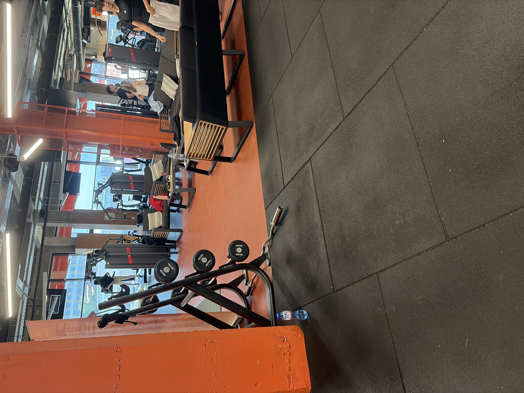 This image captures the interior of a modern gym or fitness center, characterized by an industrial aesthetic with exposed structures and pipes on the ceiling. The space features two distinct floor areas: a vibrant orange section and a dark grey, tiled rubber matting section. Large windows line one wall, revealing an urban landscape with buildings outside, suggesting it is daytime.
Several individuals are present, engaged in various gym activities. In the foreground on the orange floor, a black weight rack holds barbells with weight plates marked "25", "20", and "16" kilograms. A blue plastic water bottle is casually placed on the dark grey floor next to an orange pillar.
Further into the gym, on the orange flooring, are several pieces of exercise equipment, including treadmills and weight machines. People are seen using these machines or resting. One person in a black t-shirt and white shorts stands near a treadmill. Another person, wearing a dark t-shirt, is seated on a black couch with striped cushions, seemingly taking a break. A third person is lying on a workout bench in the mid-ground. The seating areas feature black upholstery with wooden slatted bases.
The lighting is provided by long, fluorescent-style fixtures mounted on the ceiling and walls. An orange metal mesh barrier or structure is visible in the middle ground, separating different areas. The overall impression is that of a functional, well-equipped fitness facility. Some inverted and partially obscured text is visible on an orange pillar in the lower left portion of the image.