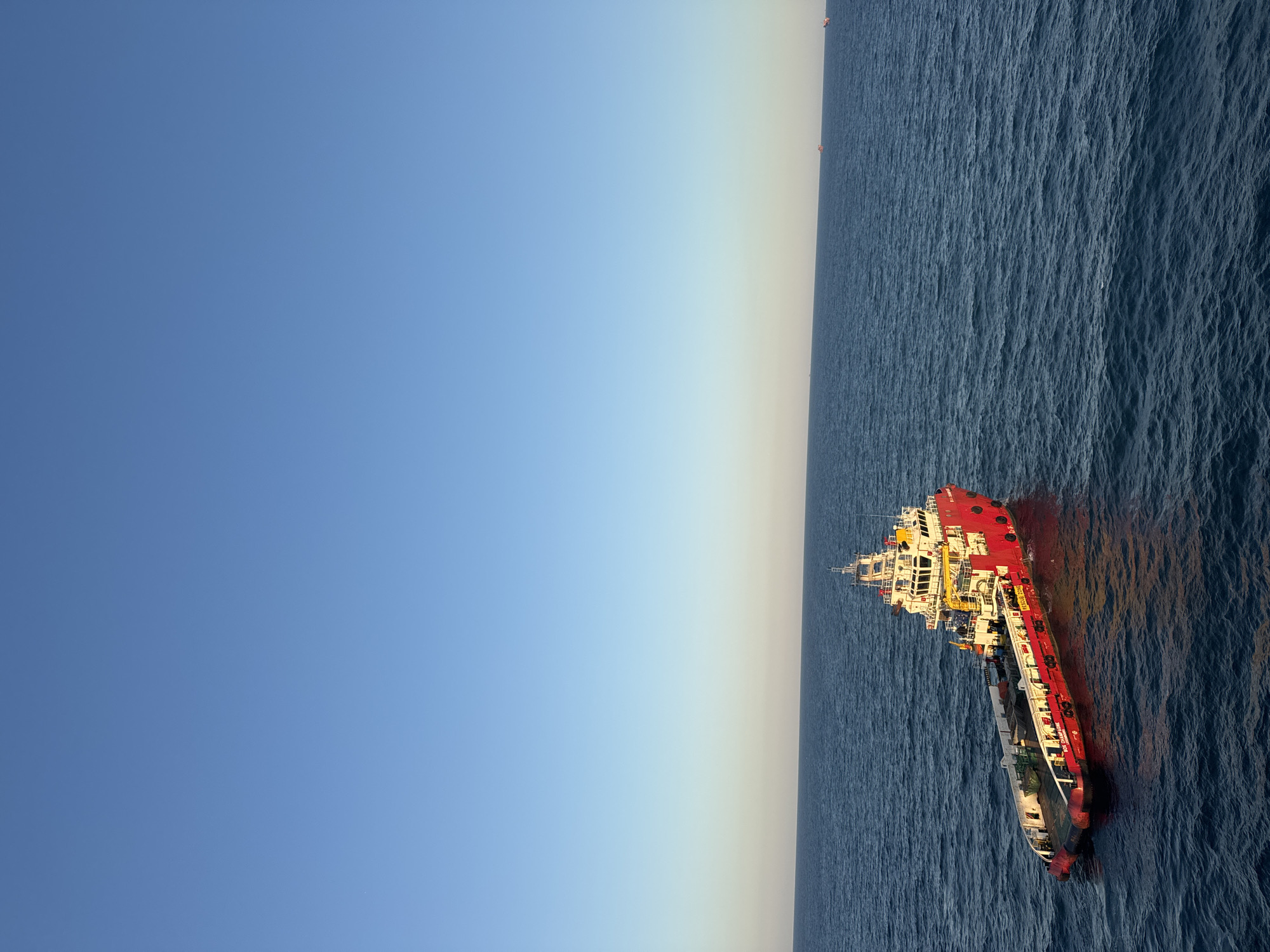 A red and white offshore supply vessel is positioned in the middle of a vast, calm blue ocean under a clear, pale blue sky. The vessel has a broad, flat deck at the stern, with various pieces of equipment and containers visible. The bow of the vessel is narrower, leading to a multi-story superstructure painted white with red accents. The ship's name, "SOL CHAMPAGNE", is visible on its side. The sun is low in the sky, casting a warm, golden light and long shadows on the water. In the distance, along the horizon, several other smaller vessels are faintly visible, suggesting this is a shipping lane or an offshore operational area. The overall scene is one of serene maritime activity.