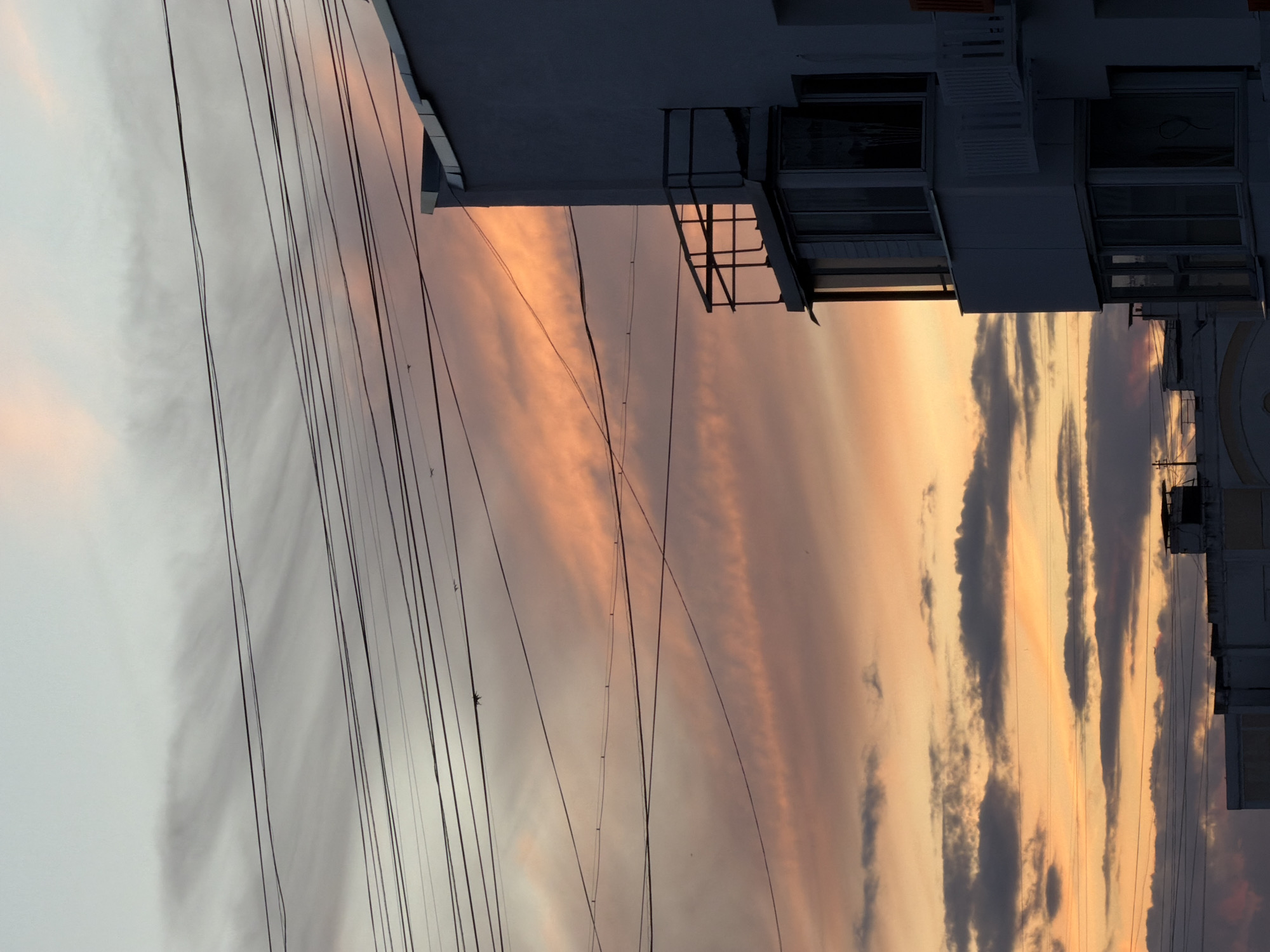 The image shows a low-angle view of a building and the sky at sunset. The sky is filled with streaks of pink, orange, and gray clouds, indicating a dramatic and colorful sunset. Several power lines crisscross the frame, obscuring parts of the sky and the building. The building appears to be modern, with multiple windows and balconies. The overall scene evokes a sense of urban twilight, with the natural beauty of the sunset contrasting with the man-made structures. The image is likely taken in Khimki, Russia, given the location context. There is no visible text in the image.