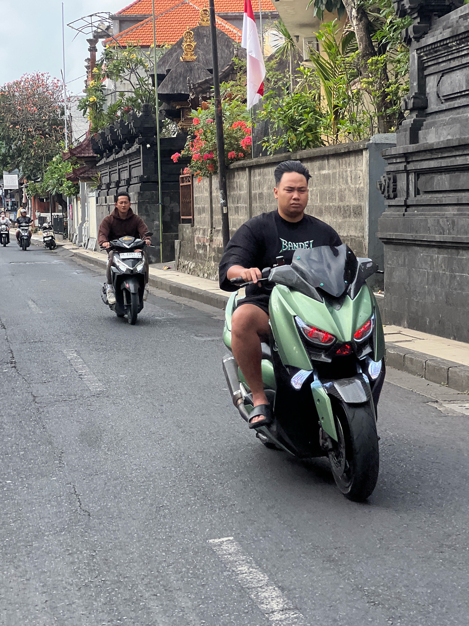 The image depicts a street scene in Kuta, Indonesia, featuring several individuals riding motorcycles. In the foreground, a man wearing a black t-shirt with the word "Bandel" and dark shorts rides a light green scooter with distinctive red taillights. To his left and further down the road, another man in a brown hooded jacket is riding a black scooter. The street appears to be paved, with some cracks and wear visible. On the right side of the street, there is a dark stone wall, and on the left, traditional Balinese architecture with orange tiled roofs and decorative elements is visible. Lush greenery and flowering plants adorn the buildings and surroundings. An Indonesian flag, red and white, flutters from a pole behind a wall. The sky is overcast, suggesting a cloudy day. Other motorcycles are visible in the background, indicating moderate traffic.