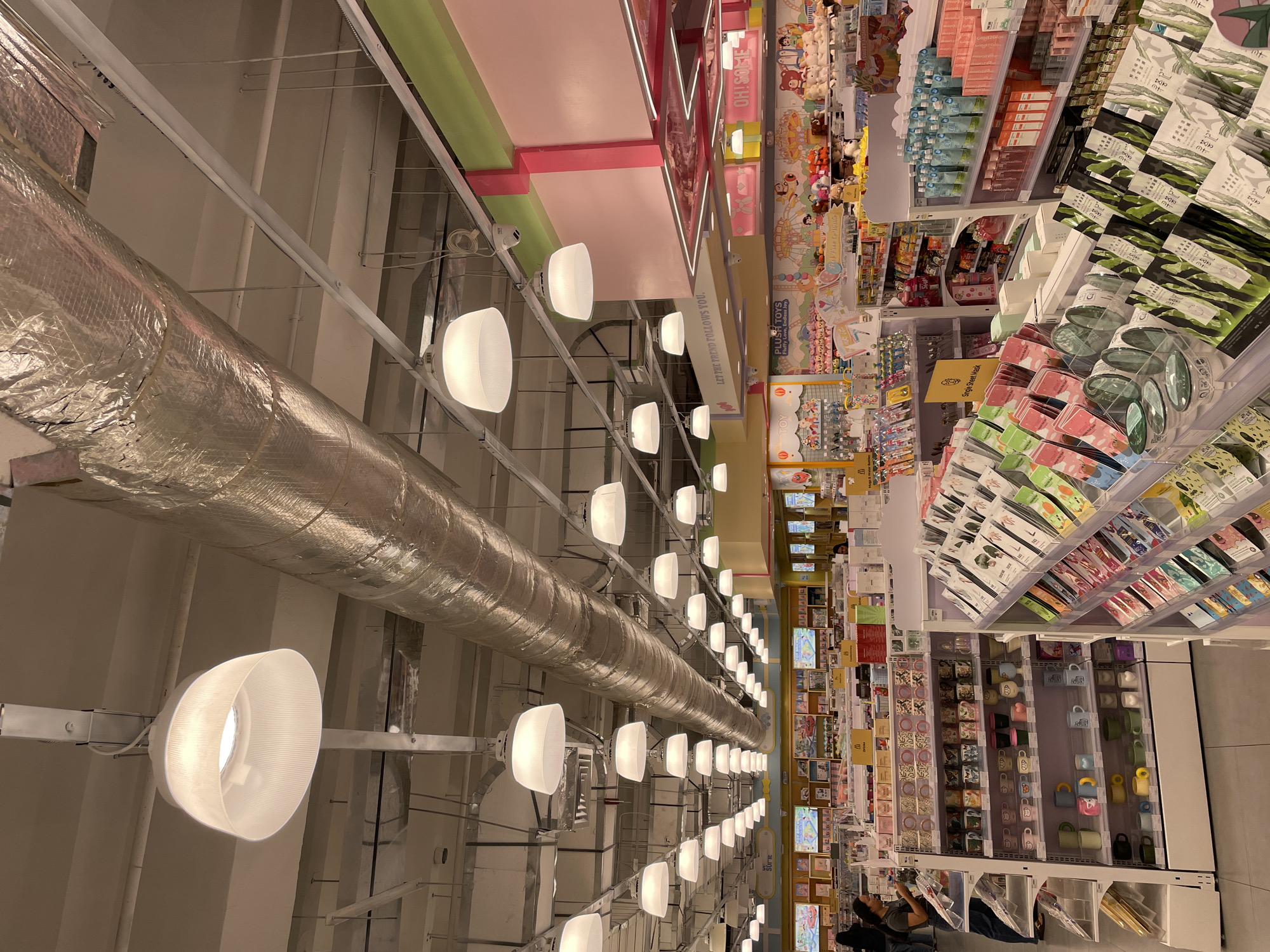 The image depicts the interior of a well-lit retail store, likely a novelty or gift shop, with numerous shelves displaying a wide array of merchandise. Overhead, a large, silver, cylindrical ventilation duct runs diagonally across the frame, supported by metal beams. Multiple white, dome-shaped pendant lights are suspended from the ceiling, illuminating the store.
The shelves are filled with various items including mugs, cups, packets of what appear to be single sheet masks, and packaged beverages or toiletries. A variety of colorful graphics and signage adorn the walls in the background, some with cartoonish characters and playful fonts, suggesting a youthful or fun-oriented product selection. Two security cameras are visible mounted on the ceiling beams.
In the foreground, two women are browsing the merchandise. One woman on the left, wearing a dark top and blue jeans, is looking at a shelf of mugs. Another woman, partially visible on the right side of the frame, is also near the shelves. The overall environment is clean, organized, and brightly lit, typical of a modern retail establishment.
The store appears to be located in Davao City, Philippines, based on the signage visible in the background which includes "OH!SOME" and "LET THE TREND FOLLOWS YOU.", and the general aesthetics common in Philippine retail spaces. The specific store is identified as an Oh!some store, a popular novelty and gift shop chain in the Philippines, with multiple branches including those in malls in Davao City.