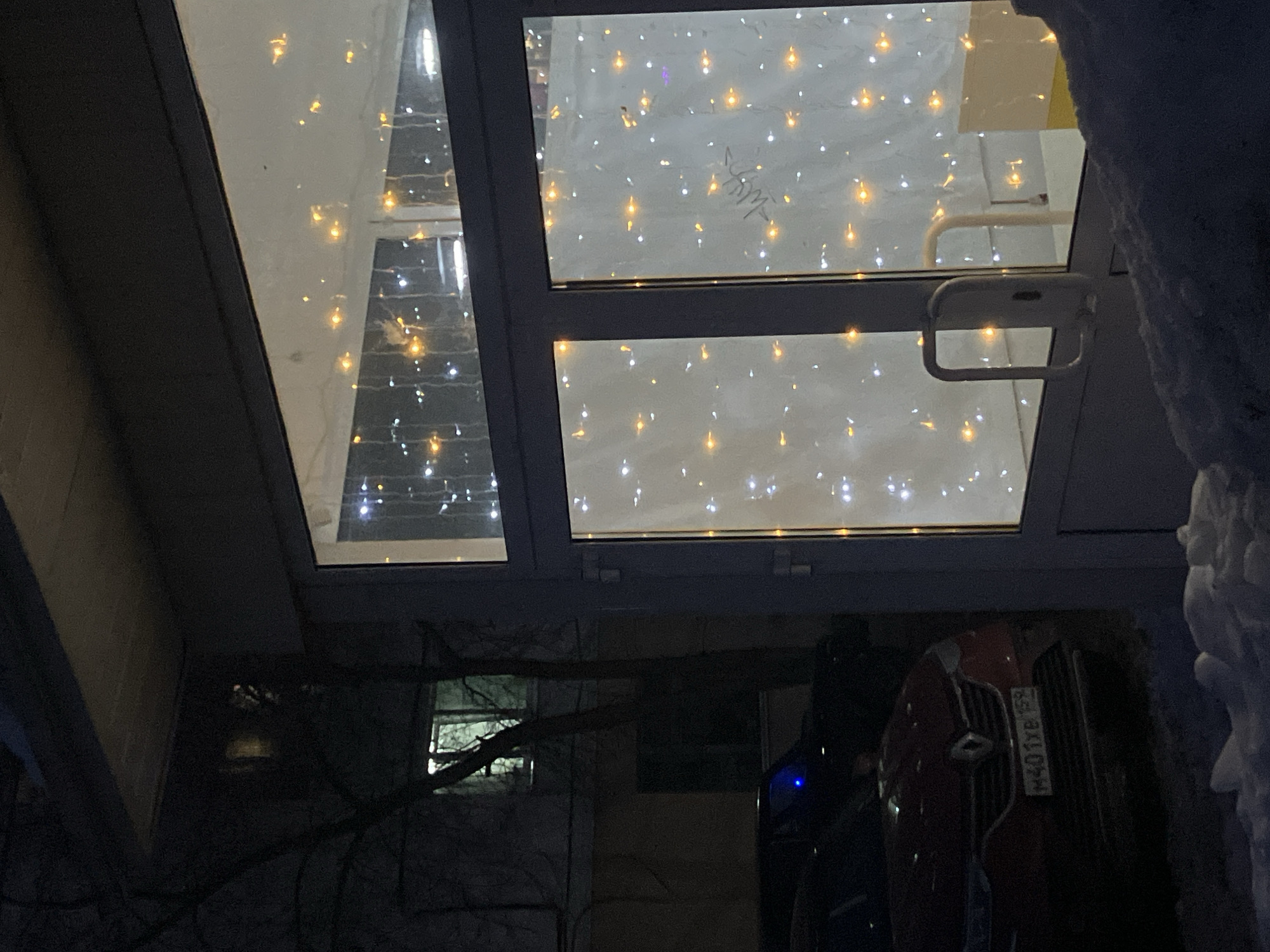The image depicts a night scene in Chaikovsky, Russia. A structure with multiple glass panels adorned with decorative lights is visible. The panels are framed in dark material. A partially obscured car, possibly red, is parked underneath. The license plate appears to read "M401YB59". A small window is located on a wall, and snow is accumulating on the right.