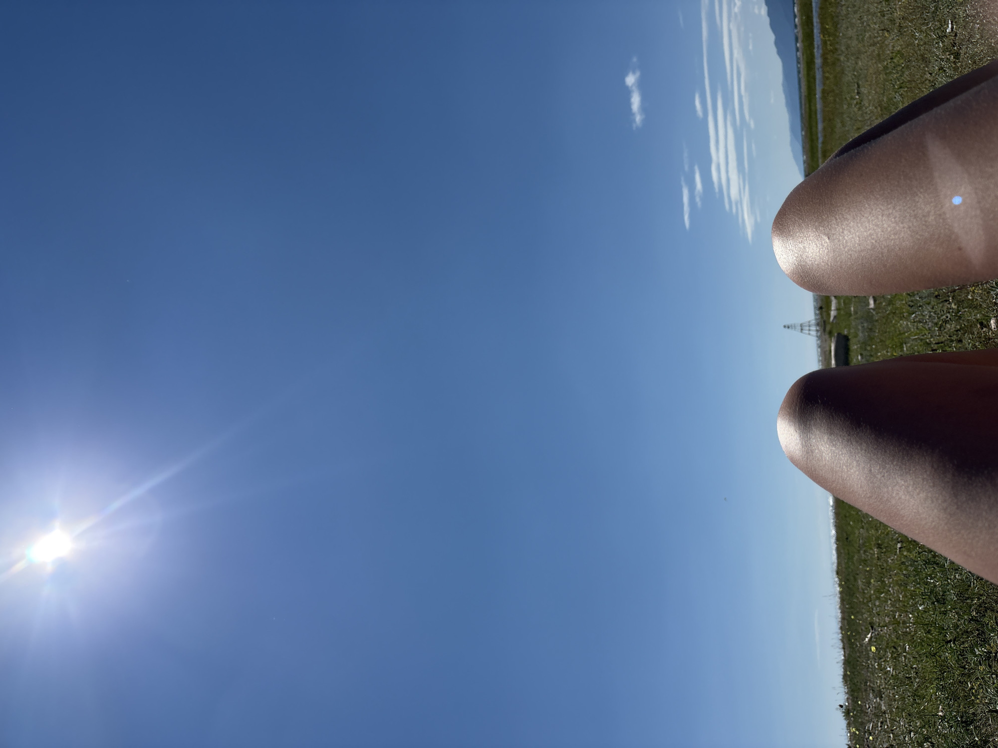 The image shows a person's legs in the foreground, with their knees visible. The legs are tanned and appear to be resting on a grassy area. The background features a clear blue sky with the sun shining brightly, creating a lens flare. A distant body of water can be seen, with hills or mountains in the far background. There is also a structure that resembles a tall, narrow tower or pylon in the middle distance. The environment appears to be a natural landscape, possibly a beach or a lakeside area. The scene suggests a warm, sunny day. The image does not contain any explicit information that would place it specifically in Bugul'deika, Russia, beyond the prompt's assertion.