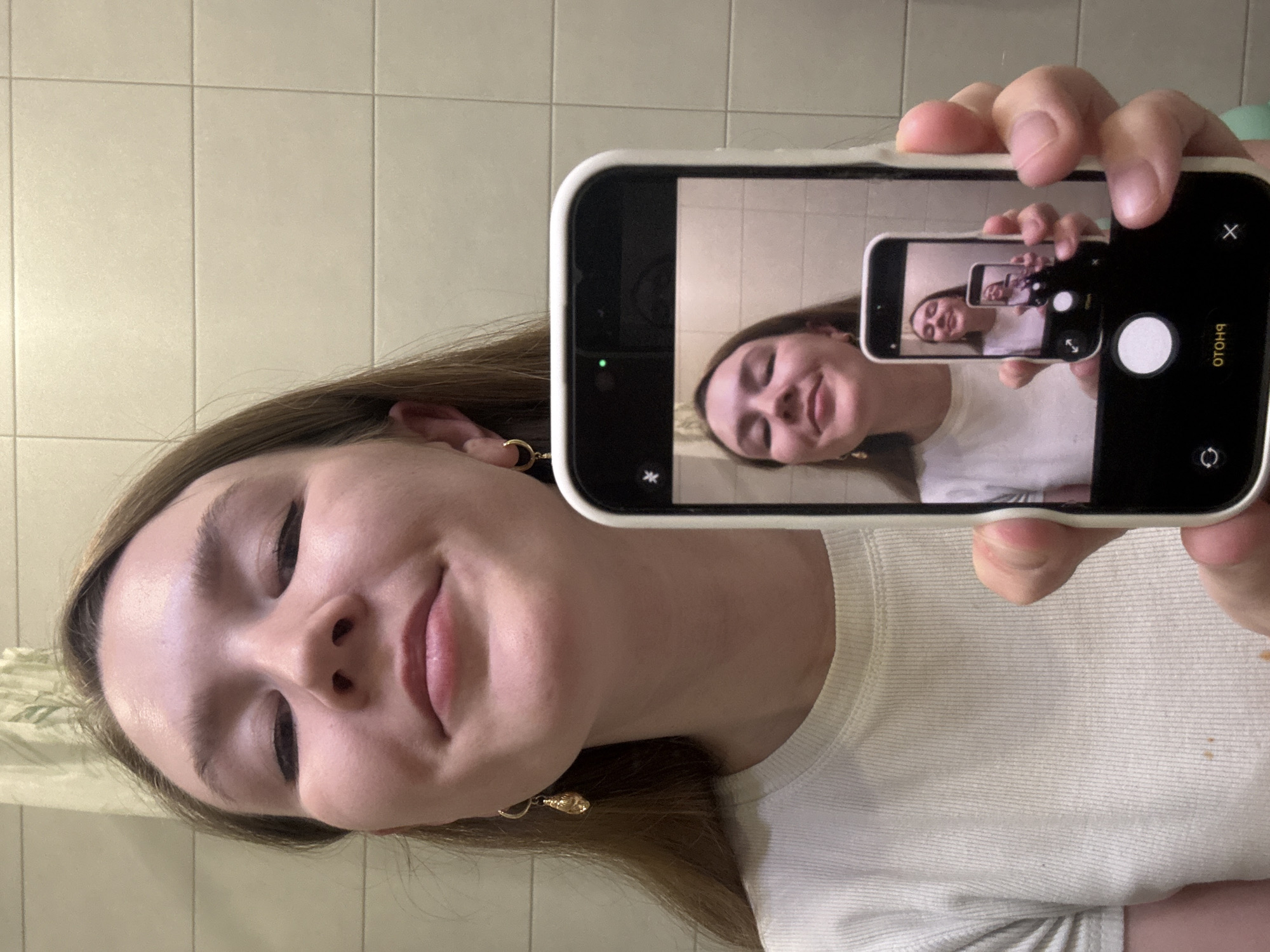 A young woman is taking a selfie in a bathroom. She holds a smartphone displaying a smaller image of herself holding a phone displaying a smaller image of herself. The background features tiled walls and a glimpse of what appears to be a shower curtain. The woman is wearing a white shirt and has dangling earrings. The smartphone in her hand is cased in white. It is not possible to determine the location as Izhevsk, Russia, from the image provided.