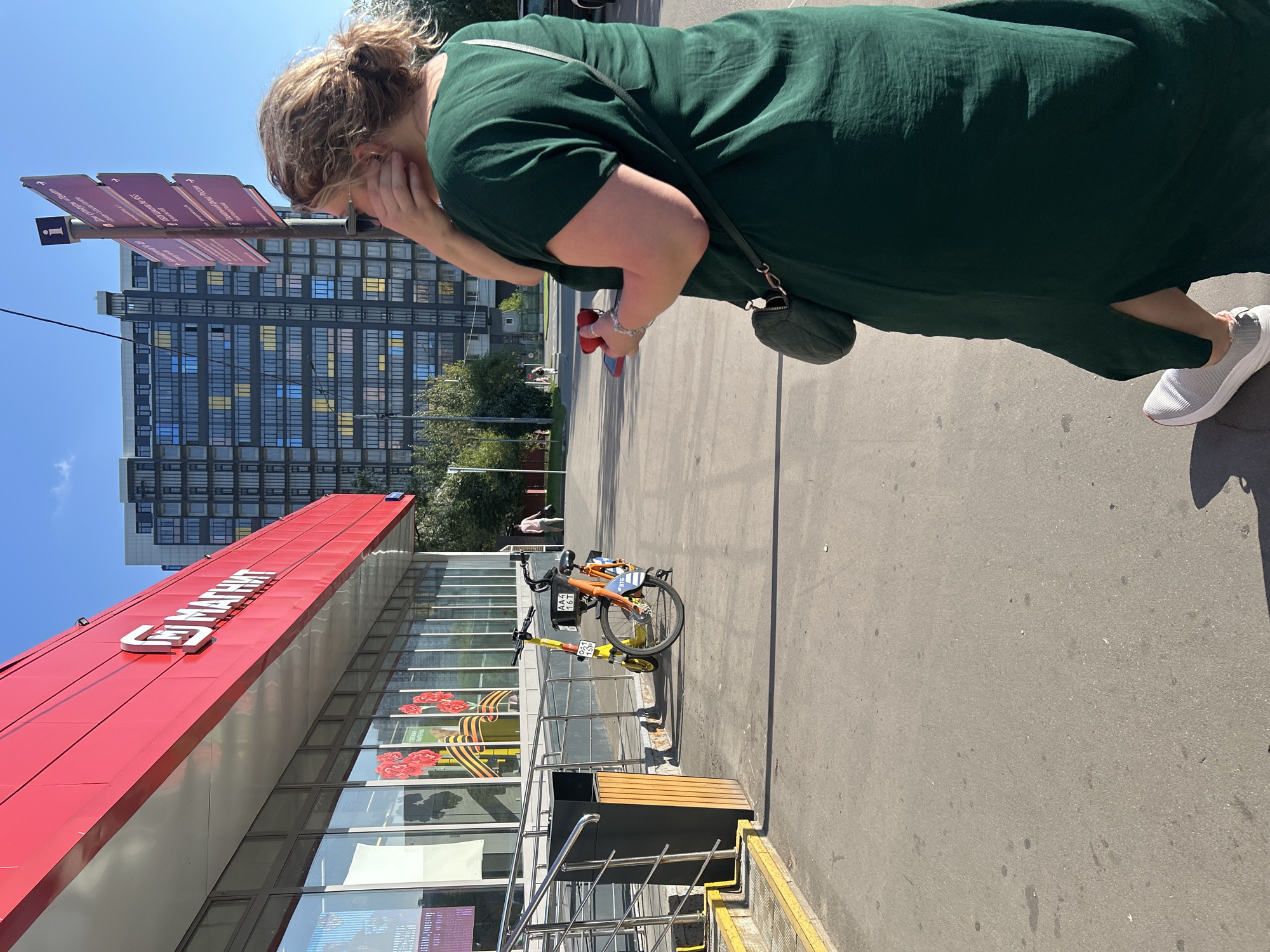 The image depicts a street scene in Moscow, Russia, with a prominent red building displaying the "Магнит" (Magnit) grocery store logo. In the foreground, a woman wearing a dark green dress and white sneakers is partially visible from behind, holding a red object in her right hand and touching her face with her left. She is carrying a small quilted purse on her left shoulder. To her left, a yellow electric scooter and an orange bicycle are parked on the sidewalk near a trash can and a ramp. In the background, a tall residential building with many windows, some of which are colored yellow and blue, dominates the skyline. A street sign with informational pictograms and text is also visible. The environment appears to be a bright, sunny day with clear blue skies.