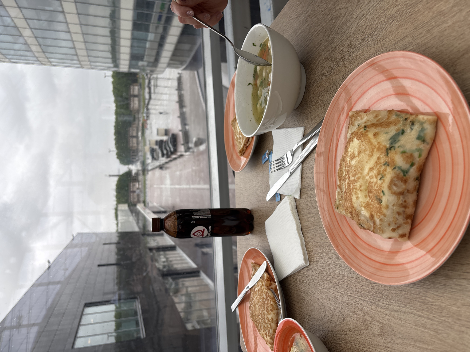 The scene depicts a meal on a wooden table in an indoor setting, likely a restaurant or cafe. A person's hand holding a spoon is visible, reaching into a white bowl of soup. Next to the bowl is a plate with what appears to be a crepe or pancake. Utensils and napkins are arranged near the plates. A bottle of dark liquid, possibly a drink, is on the table as well. Through a large window, an urban landscape is visible, including modern buildings, parked cars, and trees. The weather seems overcast. The location is in Saint Petersburg, Russia.