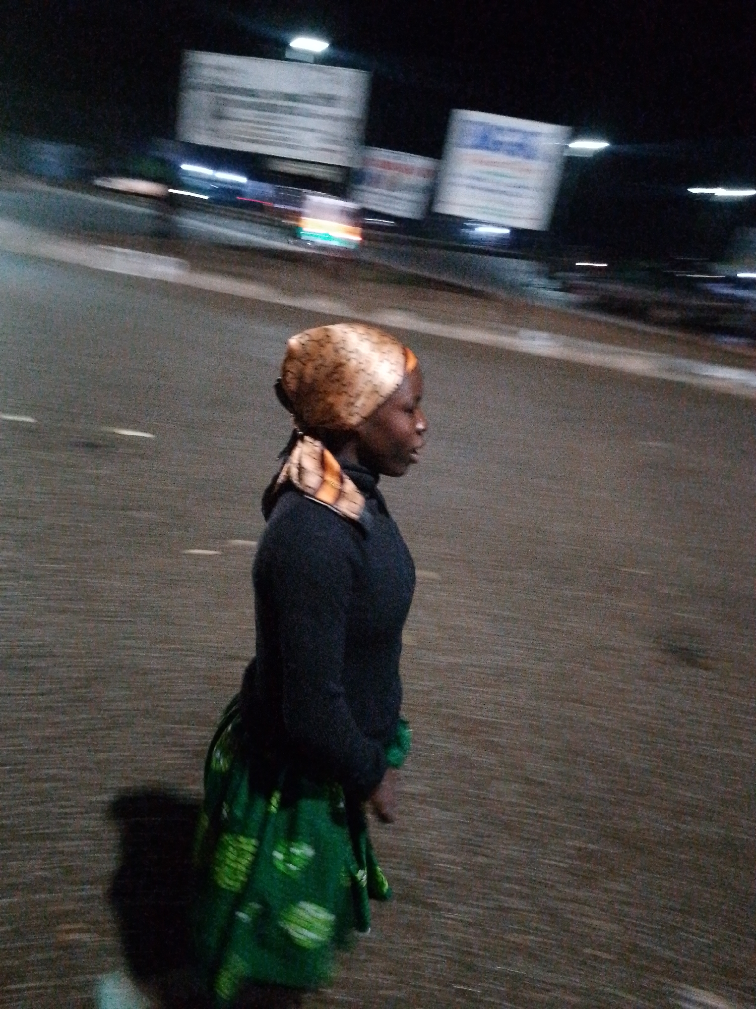 An African woman, wearing a dark long-sleeved top and a green patterned skirt, walks at night on a paved road in Bukuru, Nigeria. She has a headscarf with a gold and brown patterned fabric. The background is blurred, suggesting motion, and features streetlights and indistinct billboards, indicating an urban environment. The road surface is dark and appears to have white lane markings.