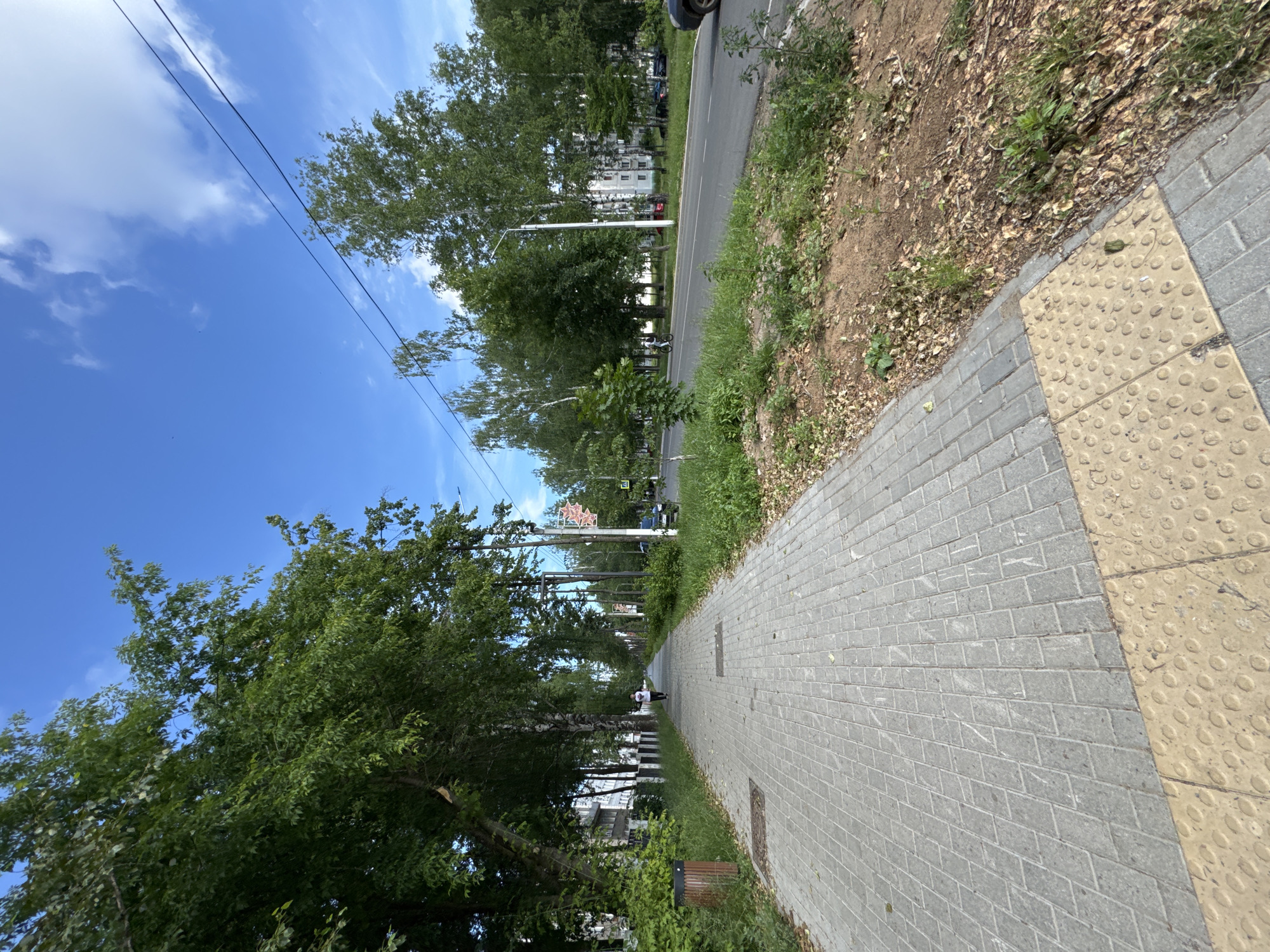The image shows a wide, paved sidewalk in Chaikovsky, Russia, lined with trees on the left and a road on the right. The sidewalk is made of gray interlocking pavers, with a tactile paving surface in yellow with raised circles at the bottom of the frame. A single person is walking away from the camera down the sidewalk. On the left, there is a trash can and a low fence, with apartment buildings visible behind the trees. On the right, there is a road with a few cars, streetlights, and more trees. The sky is blue with scattered white clouds. The scene appears to be a typical urban streetscape on a bright, clear day.