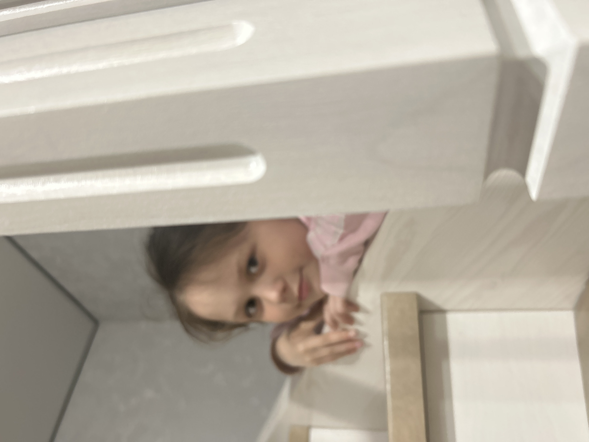 A blurred, low-angle shot features a young girl peering into the frame. She has light skin, brown hair, and wears a pink long-sleeved shirt. Her hands are visible, resting on a surface. The girl appears to be looking at the camera. The environment consists of white and grey surfaces, potentially parts of a storage unit, drawer, or cabinet. The image is blurry, and the details are difficult to discern. The location in Chaikovsky, Russia is not provided.