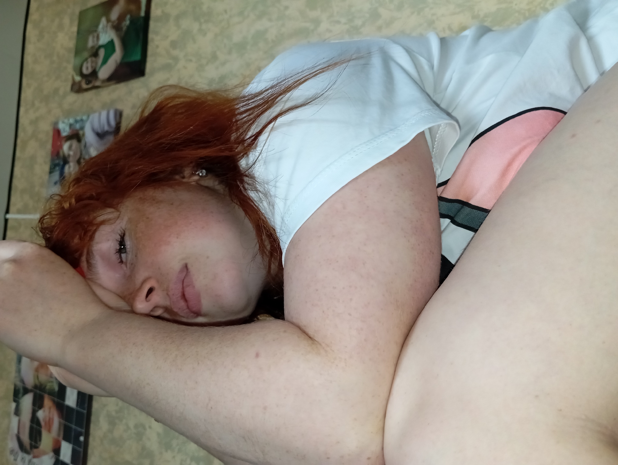 A close-up photograph shows a fair-skinned, auburn-haired woman lying on what appears to be a bed. She is wearing a white t-shirt with a pink design. Her right arm rests beneath her head, with her left arm positioned across her body. Freckles are visible on her face and arms. The background includes a wall with a light, patterned wallpaper and several small framed pictures or posters hanging on it. The location is unknown.