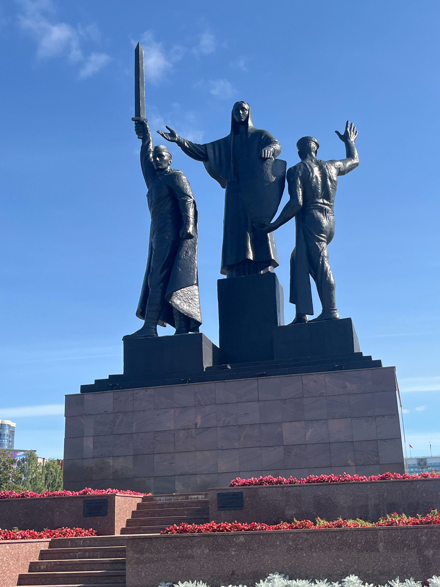 This is a photograph of the Monument to the Fighters for Soviet Power in the city of Perm, Russia. The monument is a bronze sculpture group atop a granite pedestal. The sculpture depicts three figures: a central figure in a long robe holding a shield, a figure to the left raising a sword, and a figure to the right with an outstretched hand. The figures are dark in color and appear to be made of bronze. The pedestal is tiered and covered in granite slabs. In front of the pedestal are flower beds filled with red and pink flowers, and some white foliage at the bottom. The background is a clear blue sky with a few wispy clouds. The photograph is taken from a low angle, looking up at the monument.
