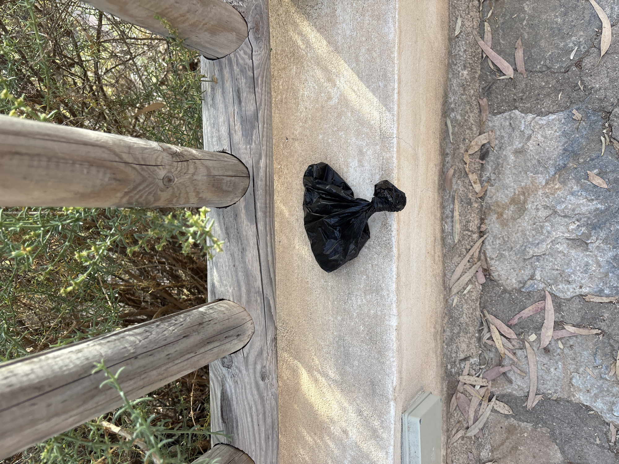 A black plastic dog waste bag is tied shut and sits on a textured, light-colored horizontal surface. Behind this surface, a wooden railing is visible, with several rough-hewn wooden posts supporting a weathered wooden beam. Beyond the railing, dense green foliage is present, characteristic of the dry Mediterranean climate found in Urbanización Cabo Roig, Spain. The ground in the foreground is paved with rough, irregularly shaped stones, some of which are covered with fallen, dry leaves. A light gray electrical or utility box is partially visible on the left side of the horizontal surface. The lighting suggests it is daytime, with dappled sunlight casting shadows on the surface where the bag rests.