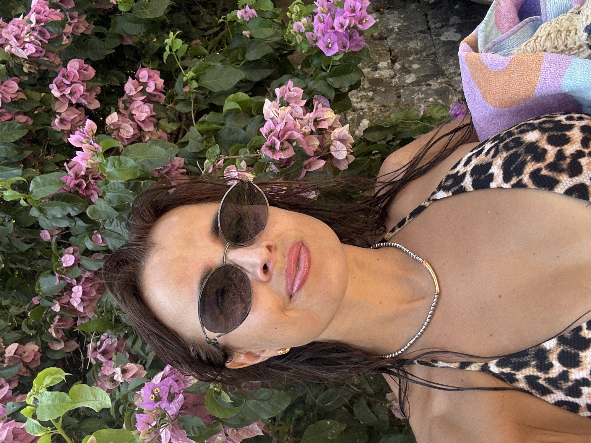 A woman with brown hair, wearing sunglasses, a bikini top, and a necklace, is pictured from the chest up. She is outdoors, with a backdrop of green leaves and pink flowers. A colorful towel is visible on her right side.