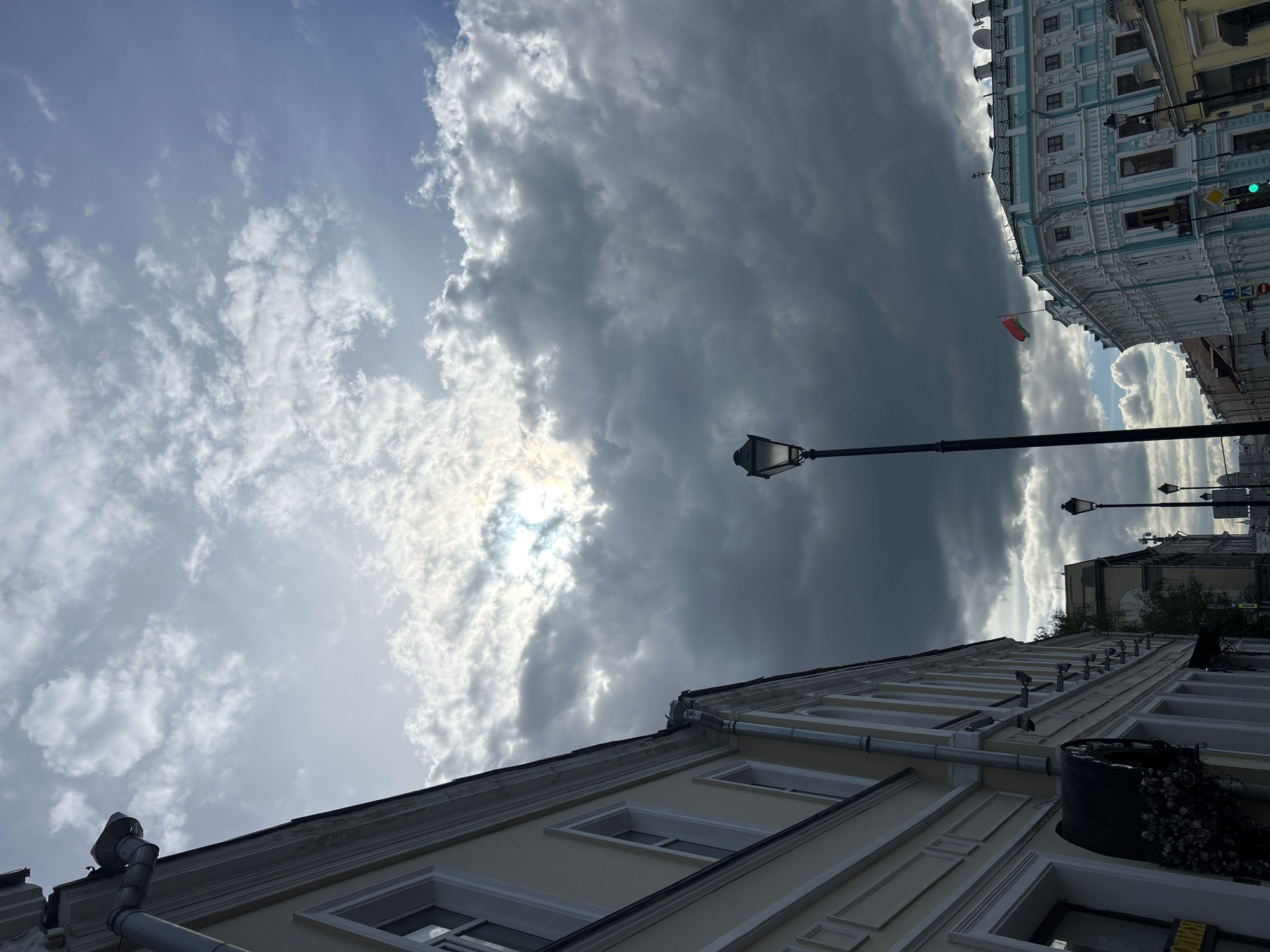 View from street level looking up at buildings and a cloudy sky. Two buildings are visible: one pale-colored with many windows and one light blue with white trim. Street lamps are positioned along the street. The sky is filled with both light and dark clouds with sunlight peeking through. A flag is seen on one of the buildings. The location is in the city of Unknown, Unknown.